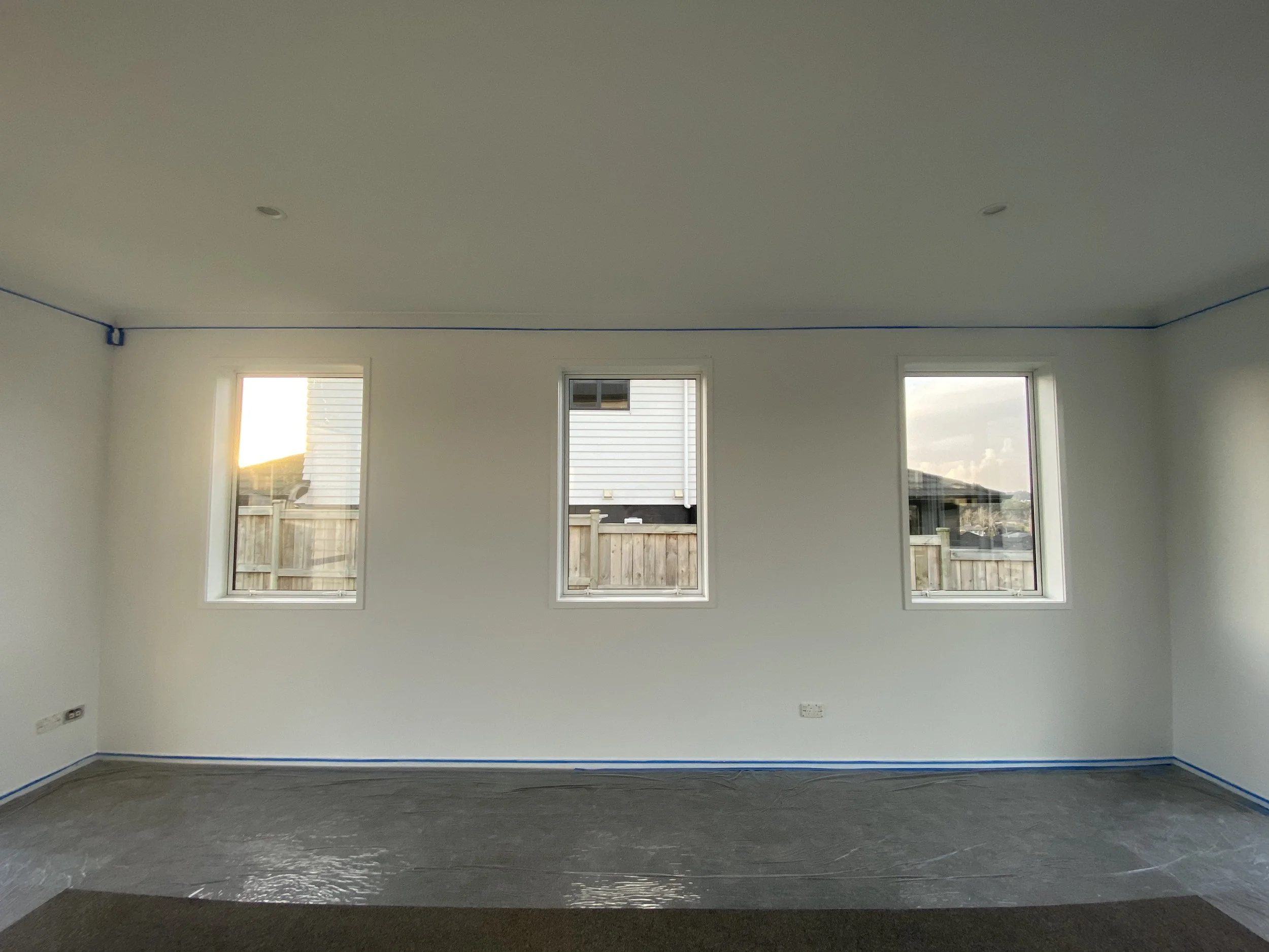 Empty room with three windows, white walls, blue baseboards, and a textured gray floor, with two ceiling lights and electrical outlets.
