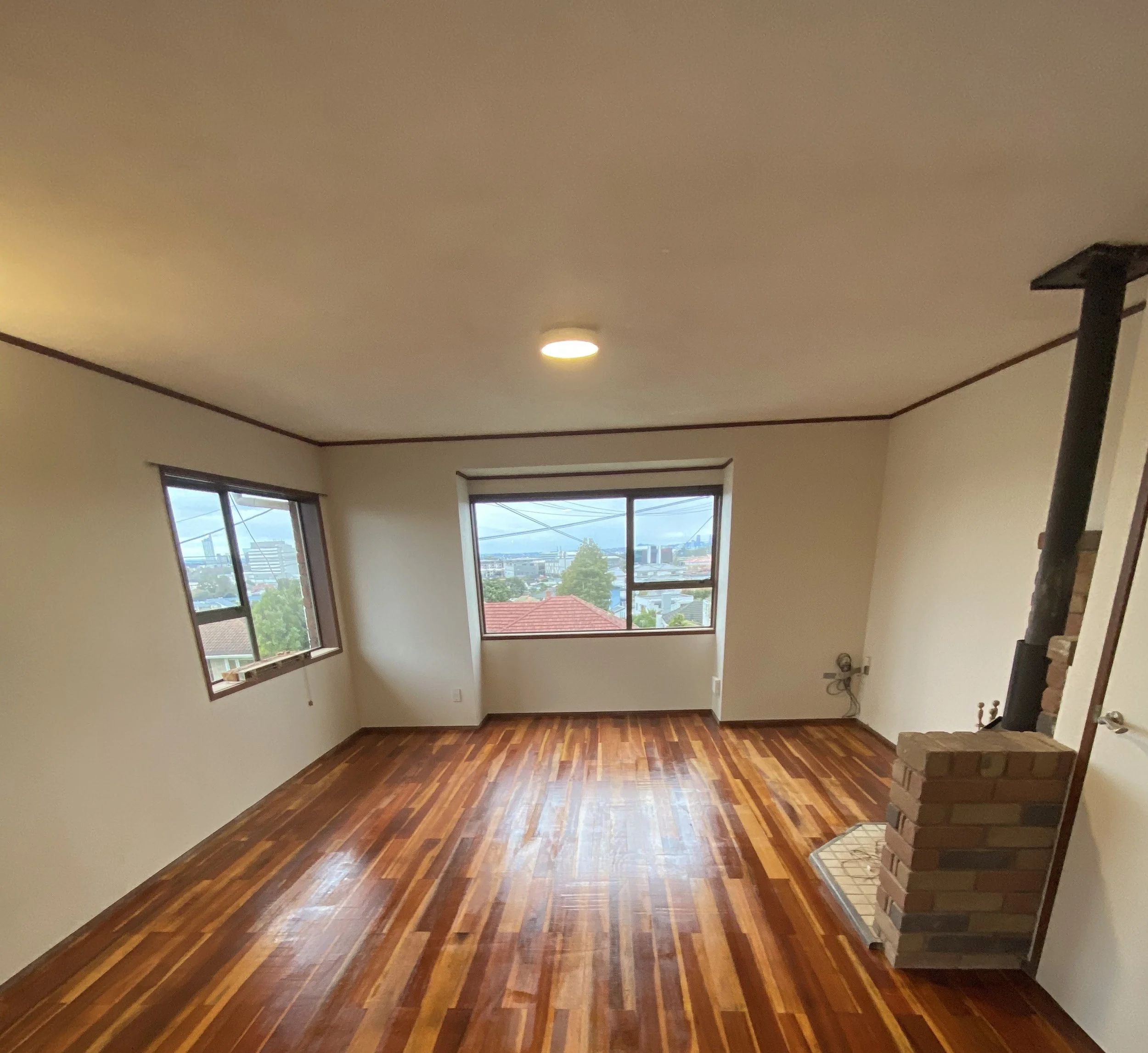 An empty living room with two large windows showing a view of a neighborhood, a hardwood floor, a ceiling light, and a brick fireplace with a timer and shelving on the right side.