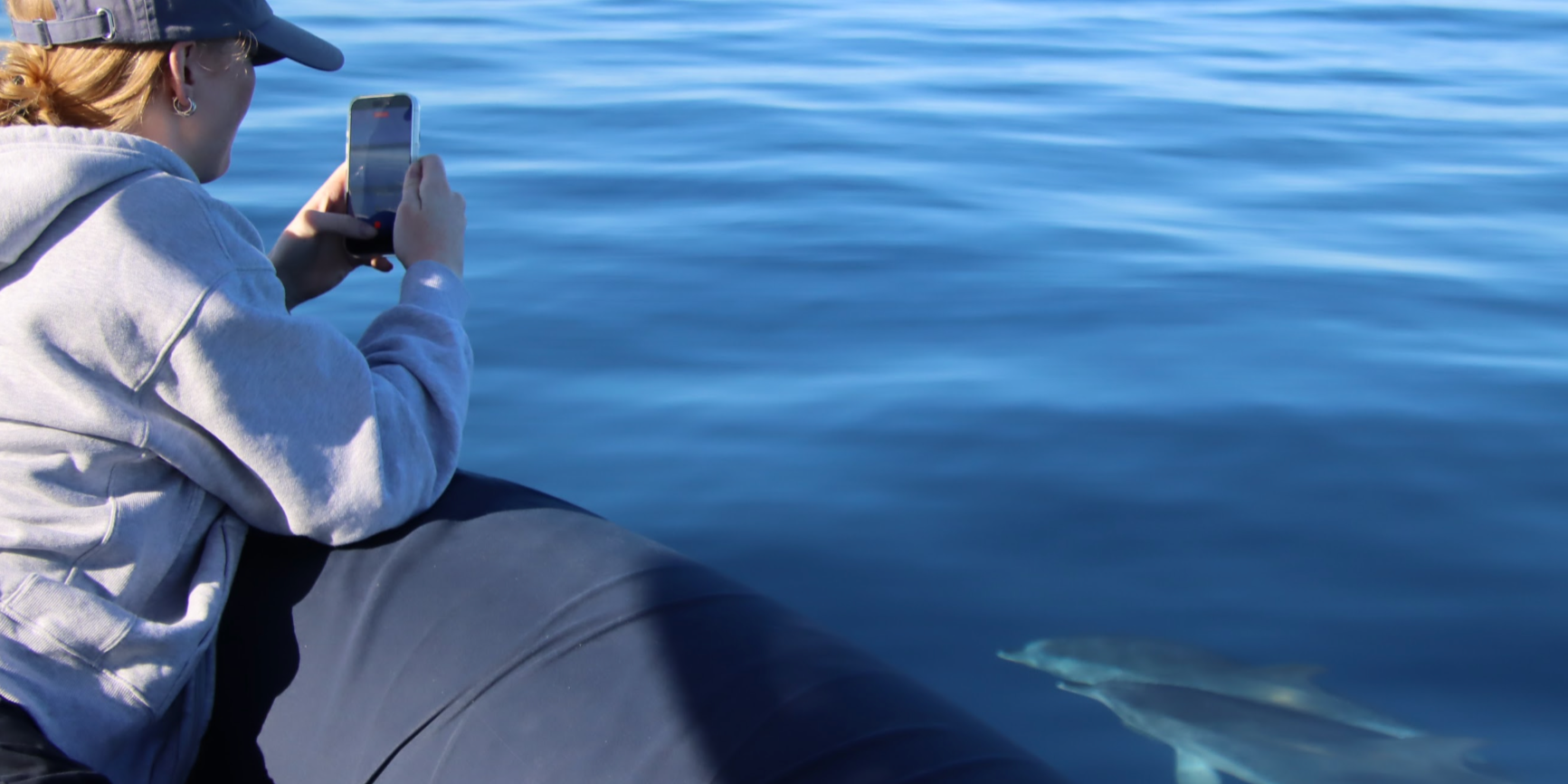 Person with red hair, wearing a cap and gray hoodie, sitting on an inflatable boat taking a photo of a dolphin swimming nearby in the ocean.