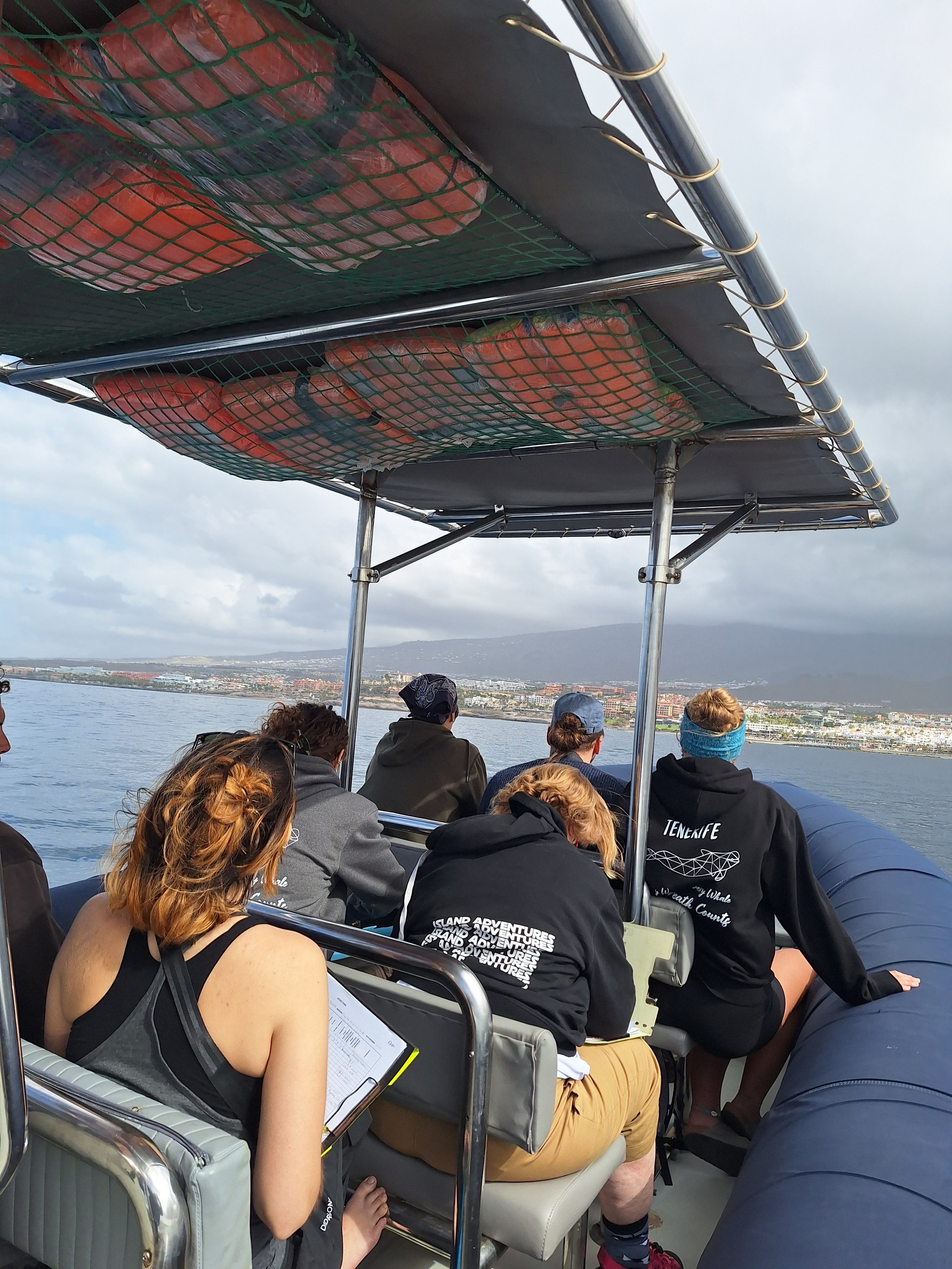 People on a boat tour looking at the water and landscape, with some taking notes and wearing casual outdoor clothing, under a canopy on a boat, with a distant shoreline and cloudy sky in the background.