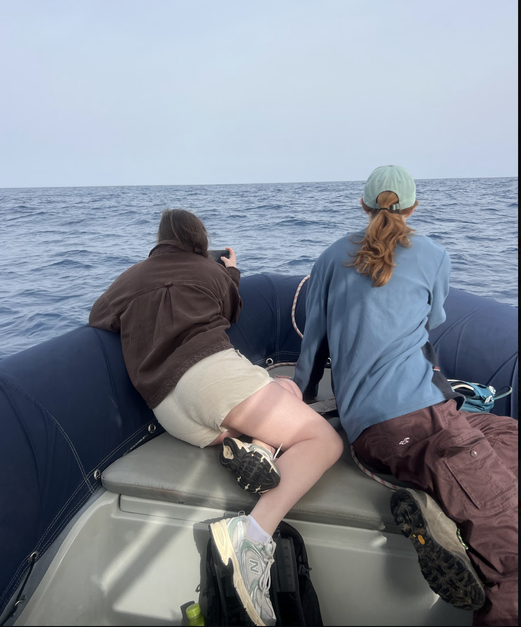 Two people on a boat looking at the ocean, one taking a photo and the other sitting with their back to the camera, wearing casual clothes and a backwards cap.