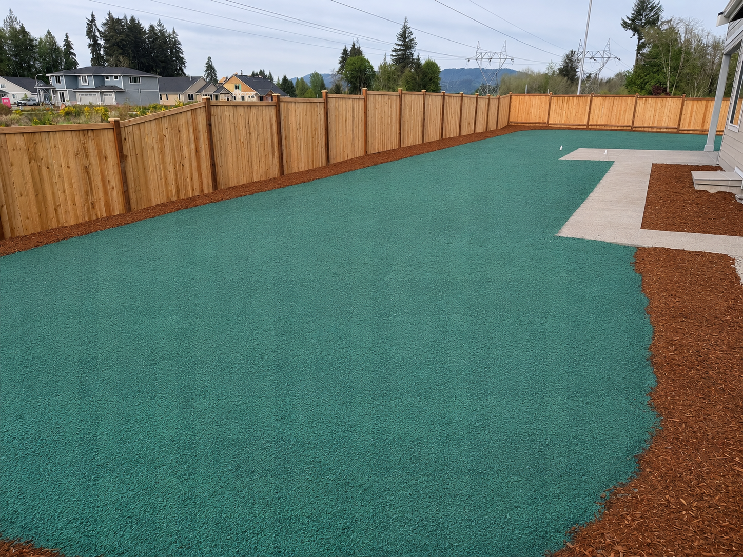 Backyard with green artificial turf, a dirt border, a concrete pathway, a wooden fence, and neighboring houses with mountains and trees in the background.
