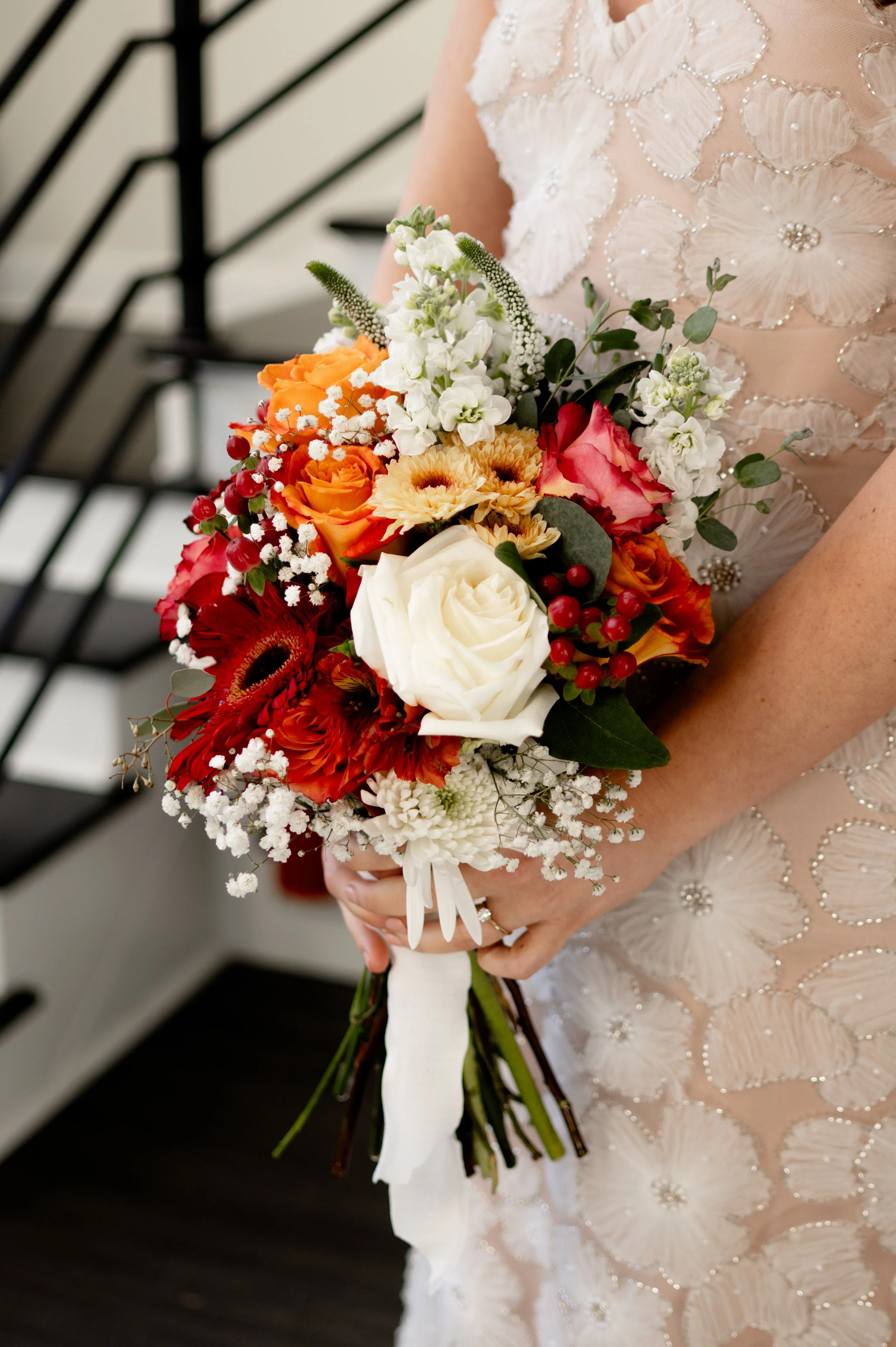 A bride holding a colorful bouquet of flowers, including white roses, orange roses, red flowers, white baby's breath, and greenery, in front of her wedding dress with intricate floral embellishments.