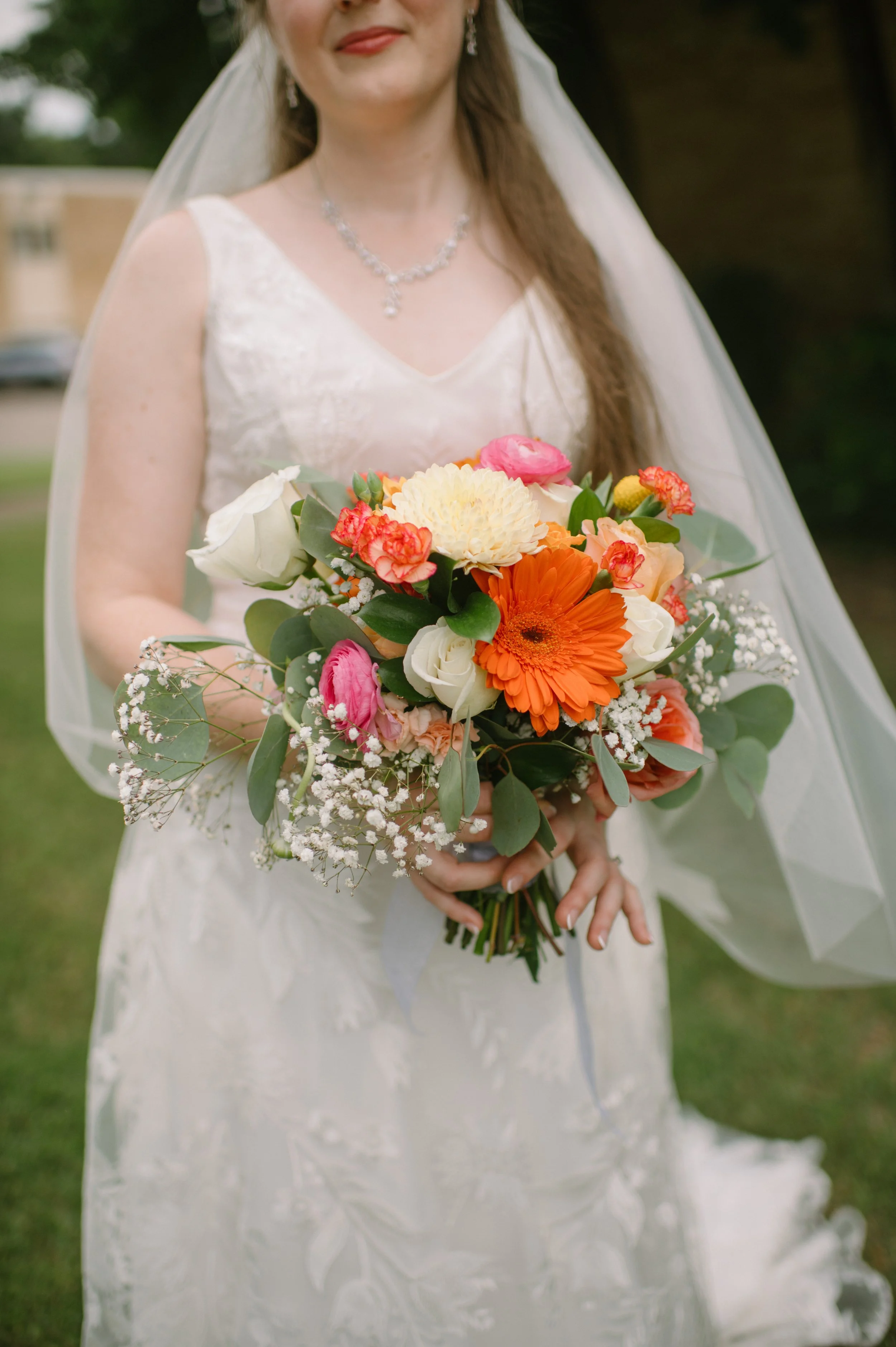 A bride in a white wedding dress holding a vibrant bouquet of mixed flowers, including orange, pink, white, cream, and green foliage, outdoors with a background of green grass and trees.