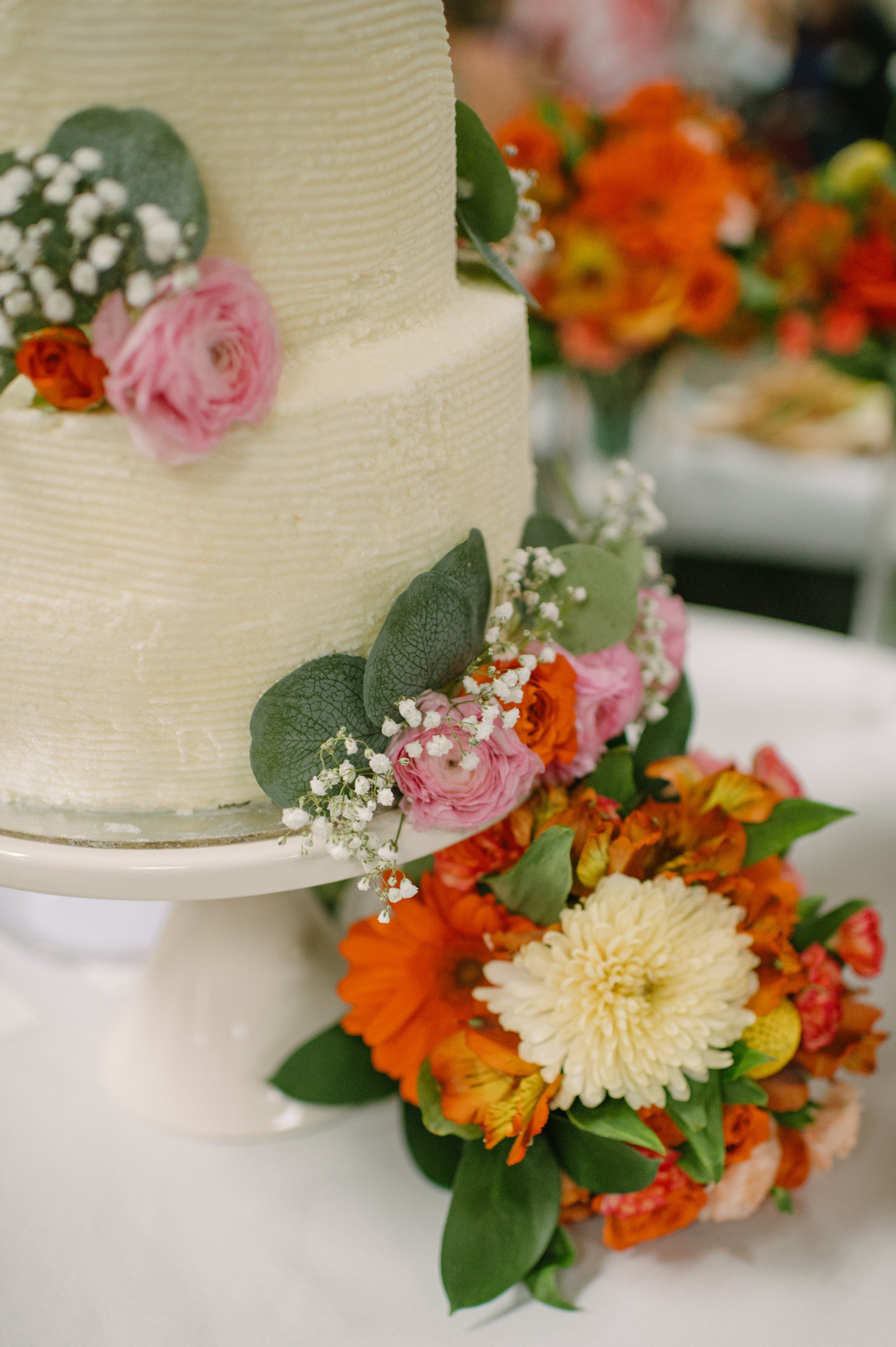 Close-up of a wedding cake decorated with pink, orange, and white flowers, green leaves, and baby's breath, with additional floral arrangements in the background.