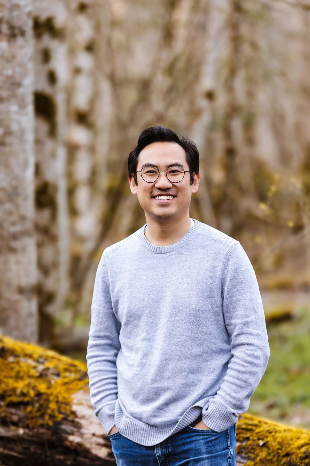 A smiling man with glasses and dark hair stands outdoors among trees with blurred branches in the background. He is wearing a light gray sweater and blue jeans.