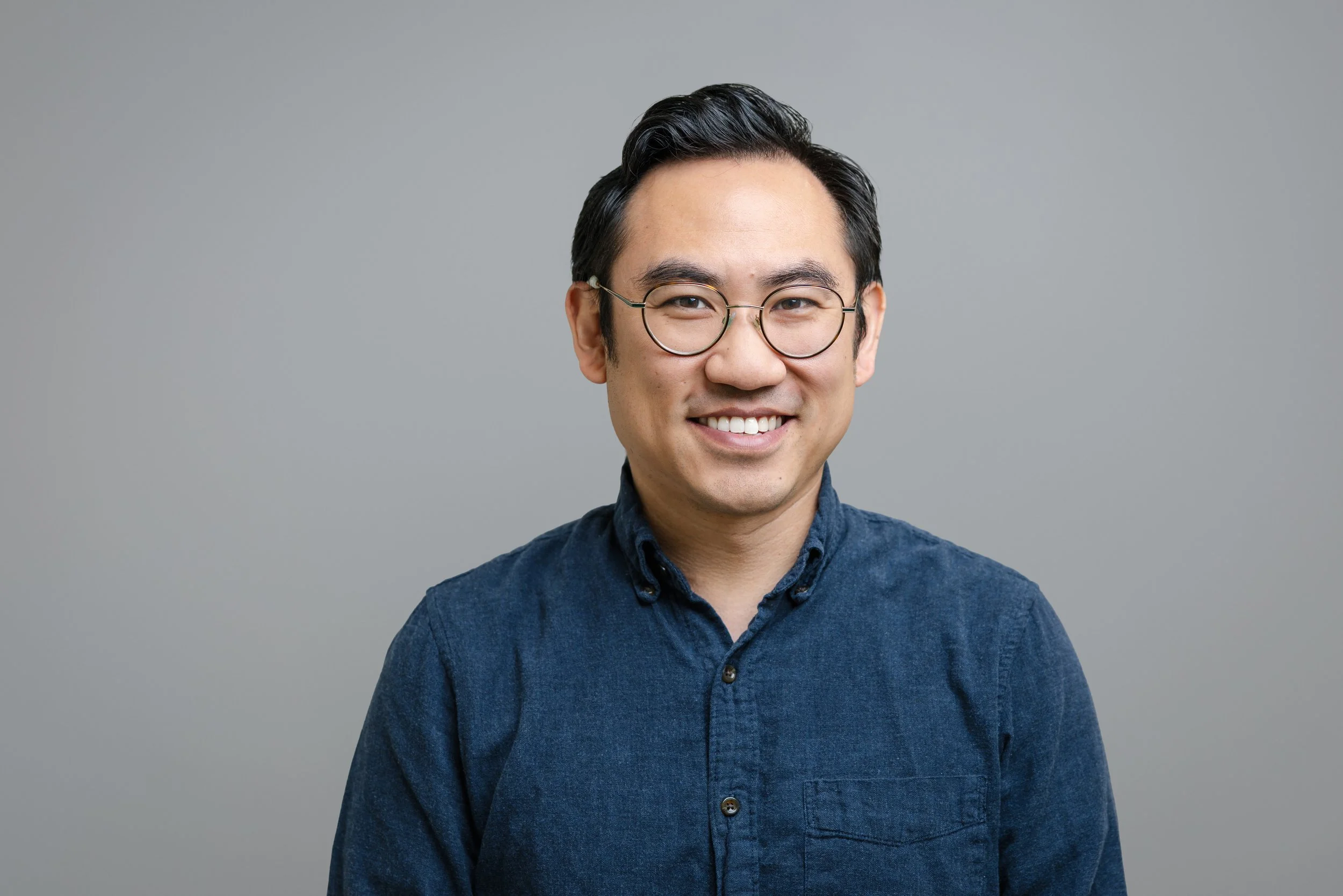 Portrait of a young man with black hair and glasses, smiling, wearing a blue denim shirt against a plain gray background.