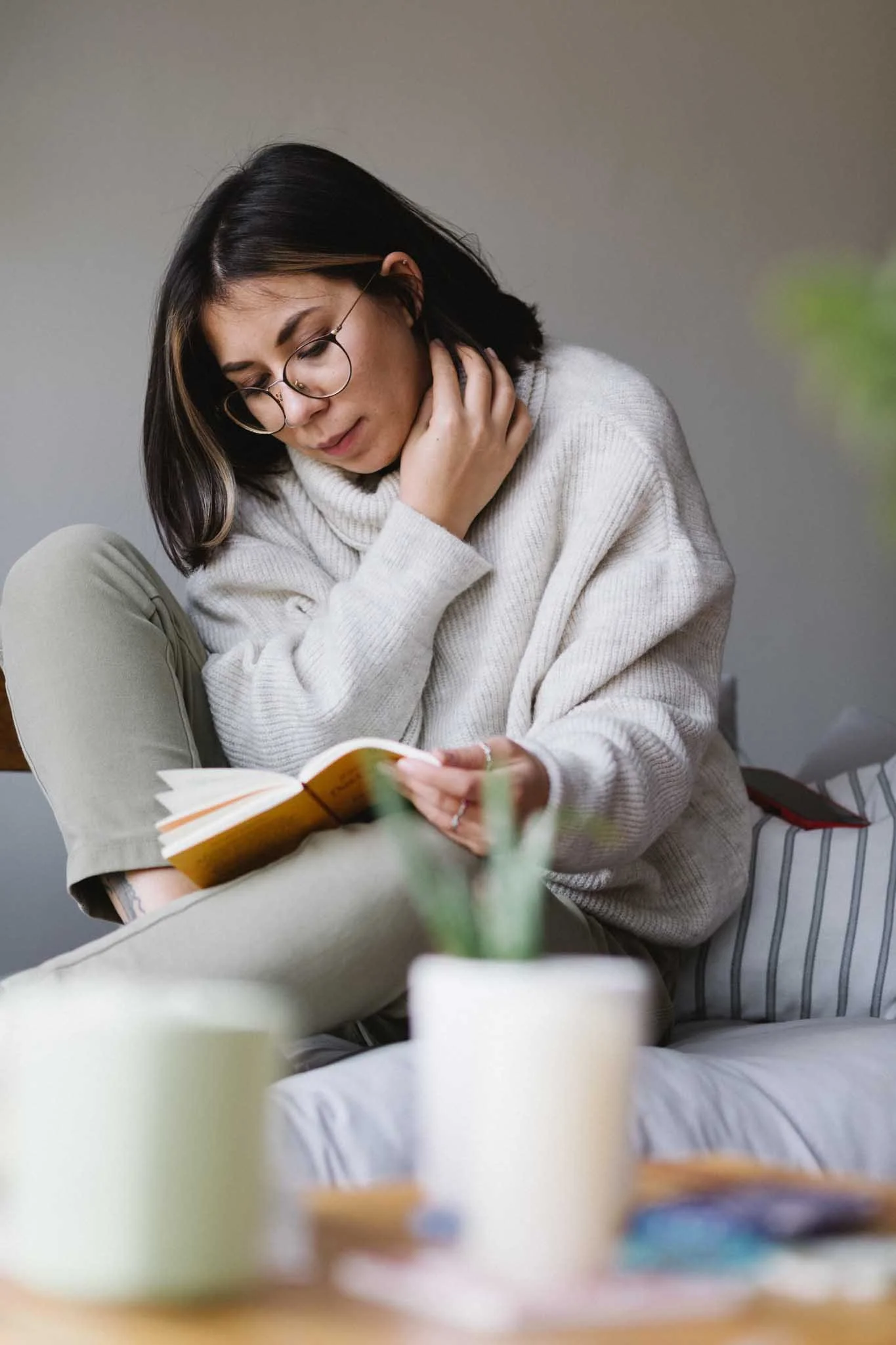 A woman with shoulder-length dark hair, glasses, and a white sweater sits on a couch, reading a book. The background is plain and neutral, and in the foreground, out-of-focus cups and a potted plant are visible.