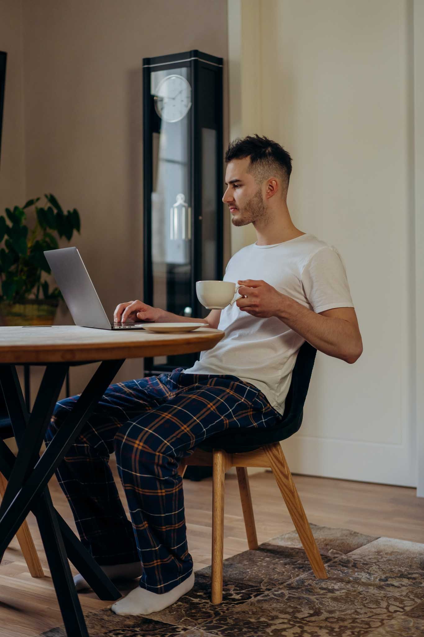 A young man wearing pajamas and white slippers sitting at a wooden table, working on a silver laptop, holding a white mug, in a cozy room with wood flooring, a plant, and a vintage grandfather clock in the background.