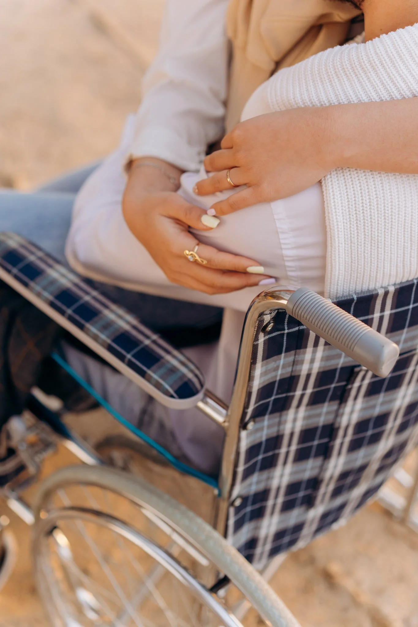 Close-up of a person's hand with rings grasping the armrest of a wheelchair with a checkered fabric seat and backrest on a dirt ground outdoors.
