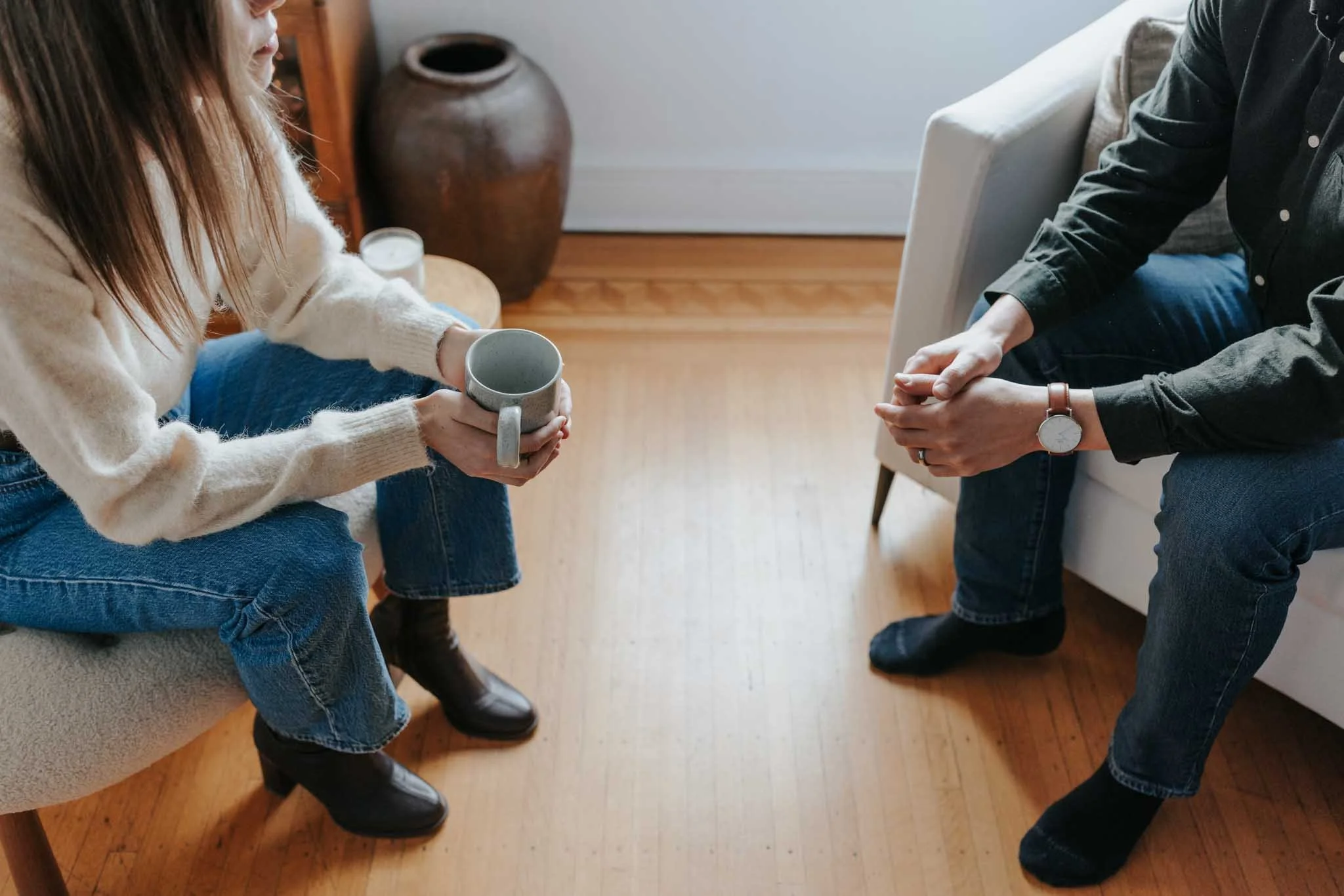 Two people sitting across from each other with their hands clasped, holding coffee mugs. One person is wearing a beige sweater and jeans, and the other is wearing a black shirt and a watch. They are in a cozy room with wooden flooring and a ceramic vase in the background.
