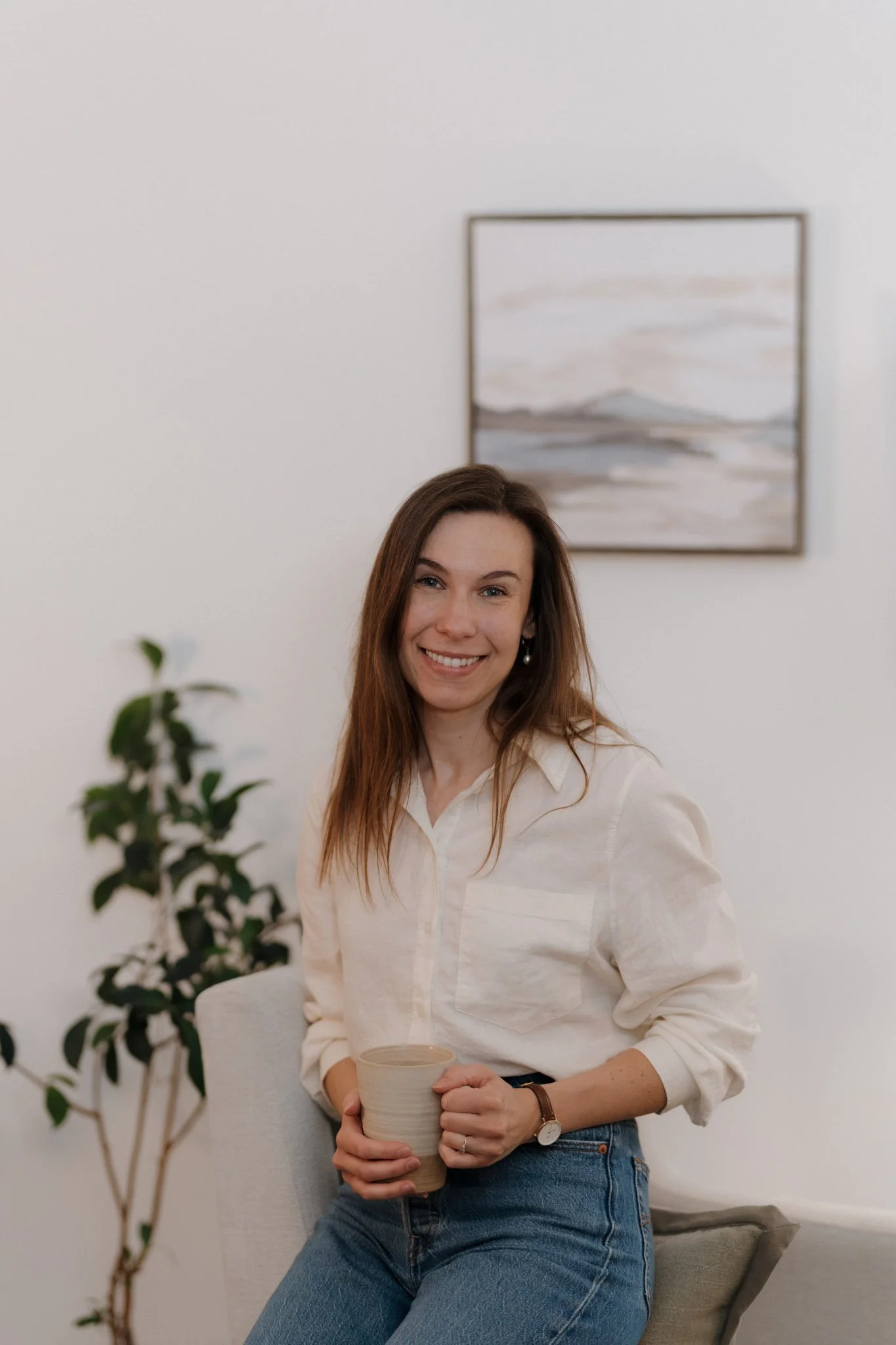A woman with long brown hair smiling and holding a beige mug, sitting on a light-colored chair in a room with a white wall, a plant, and a framed abstract painting in the background.