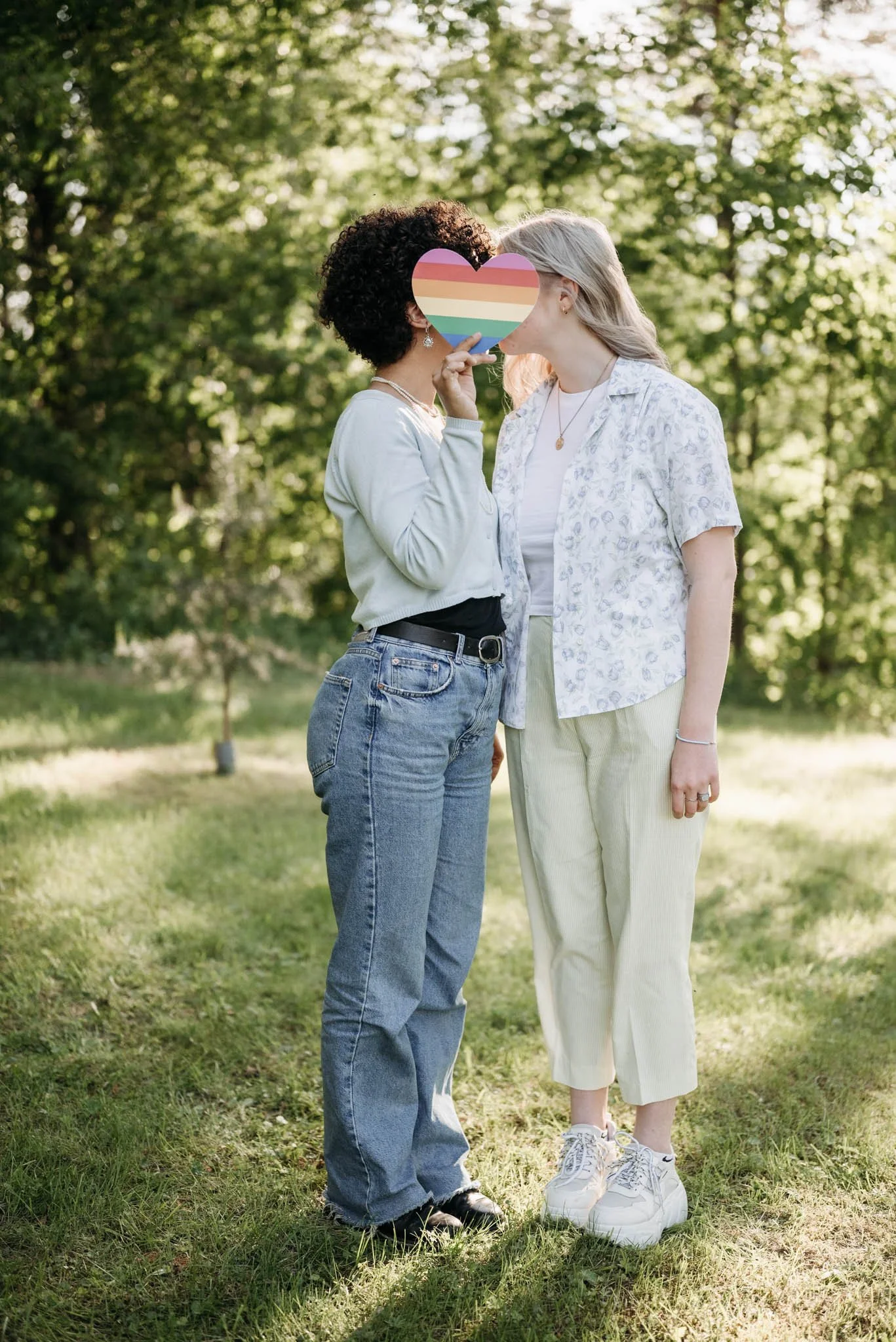 Two women standing outdoors in a park with green trees in the background, holding a rainbow-colored heart-shaped sign in front of their faces, kissing.