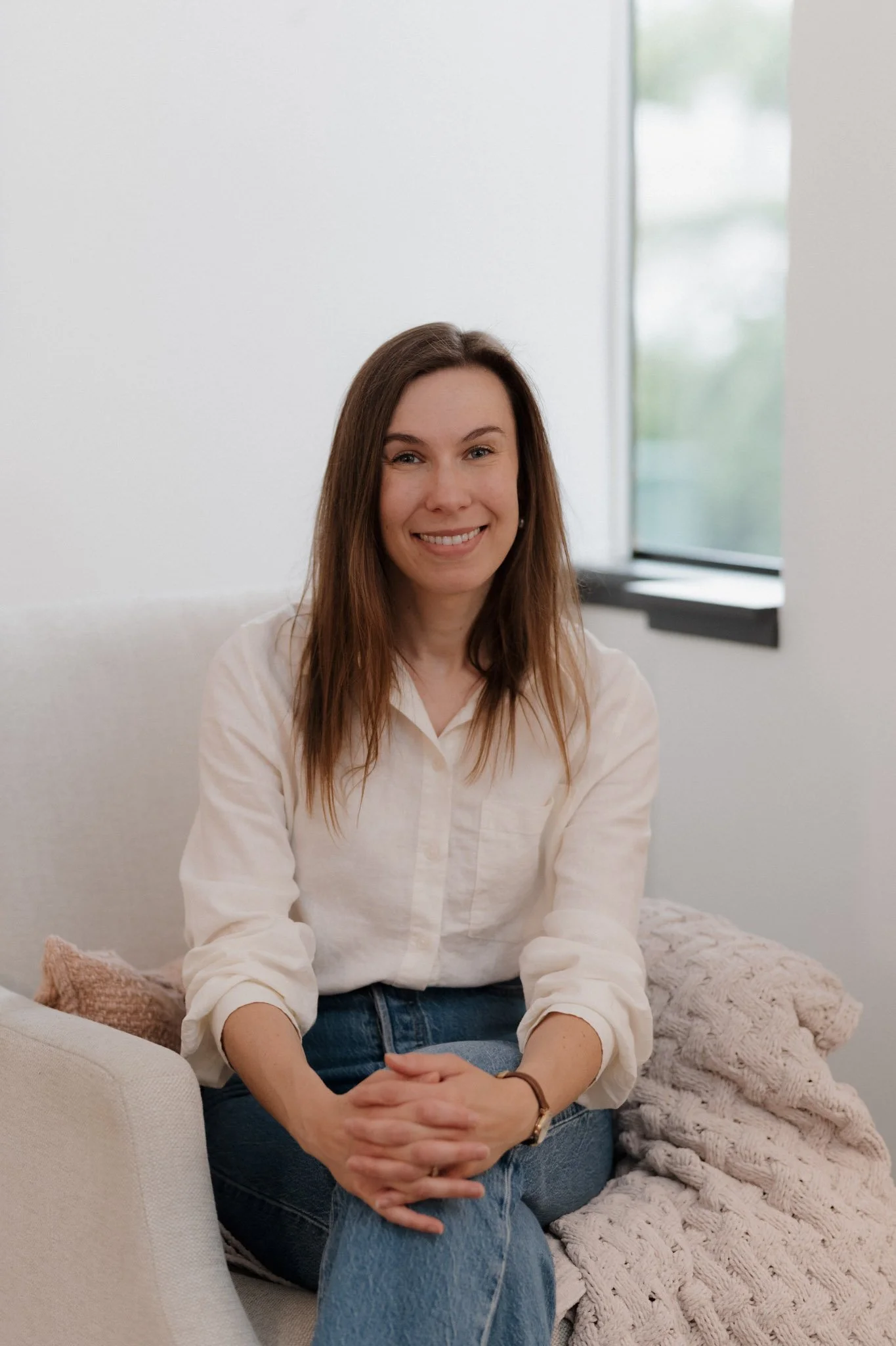 A woman with brown hair, wearing a light-colored button-up shirt and jeans, sitting on a sofa near a window in a bright room, smiling at the camera.
