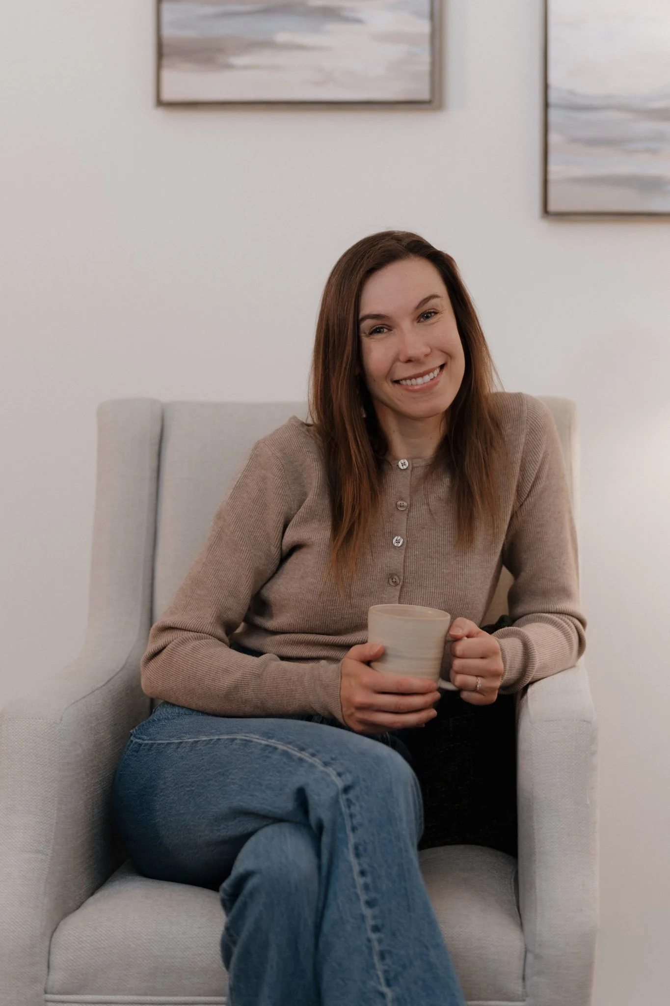 A woman with long brown hair sitting on a light-colored armchair, holding a beige mug, smiling, in a room with abstract paintings on the wall.