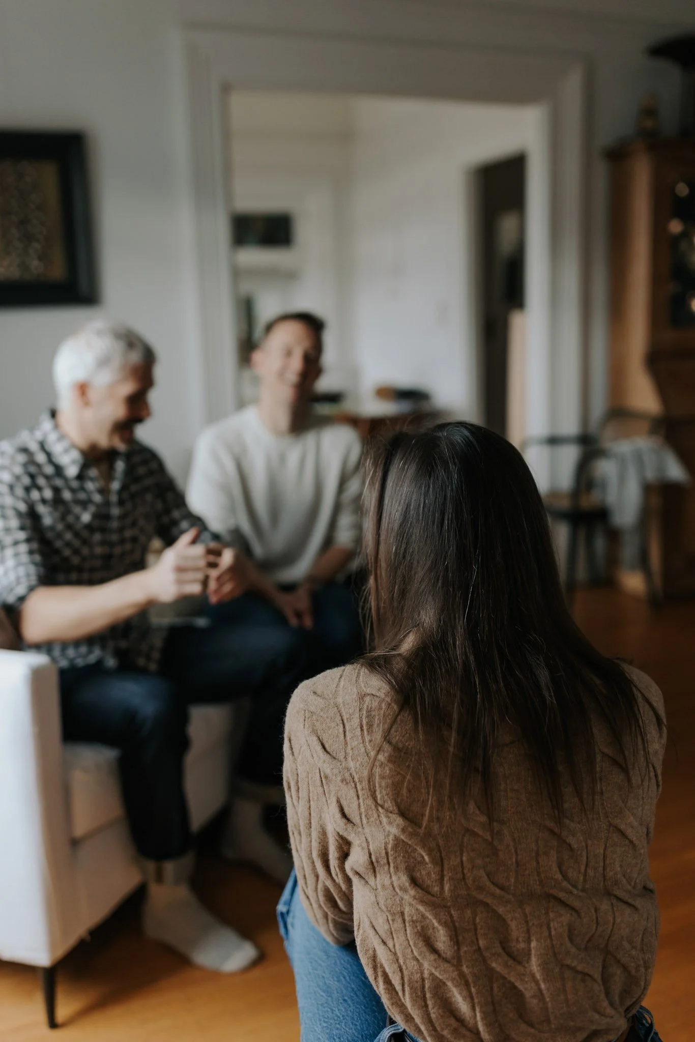 People gathered in a living room, sitting and standing while engaged in conversation. The focus is on a woman with long dark hair and a brown sweater, with two other women in the background, one with white hair and a checkered shirt, smiling, and another smiling woman with dark hair tied back, in a white top.