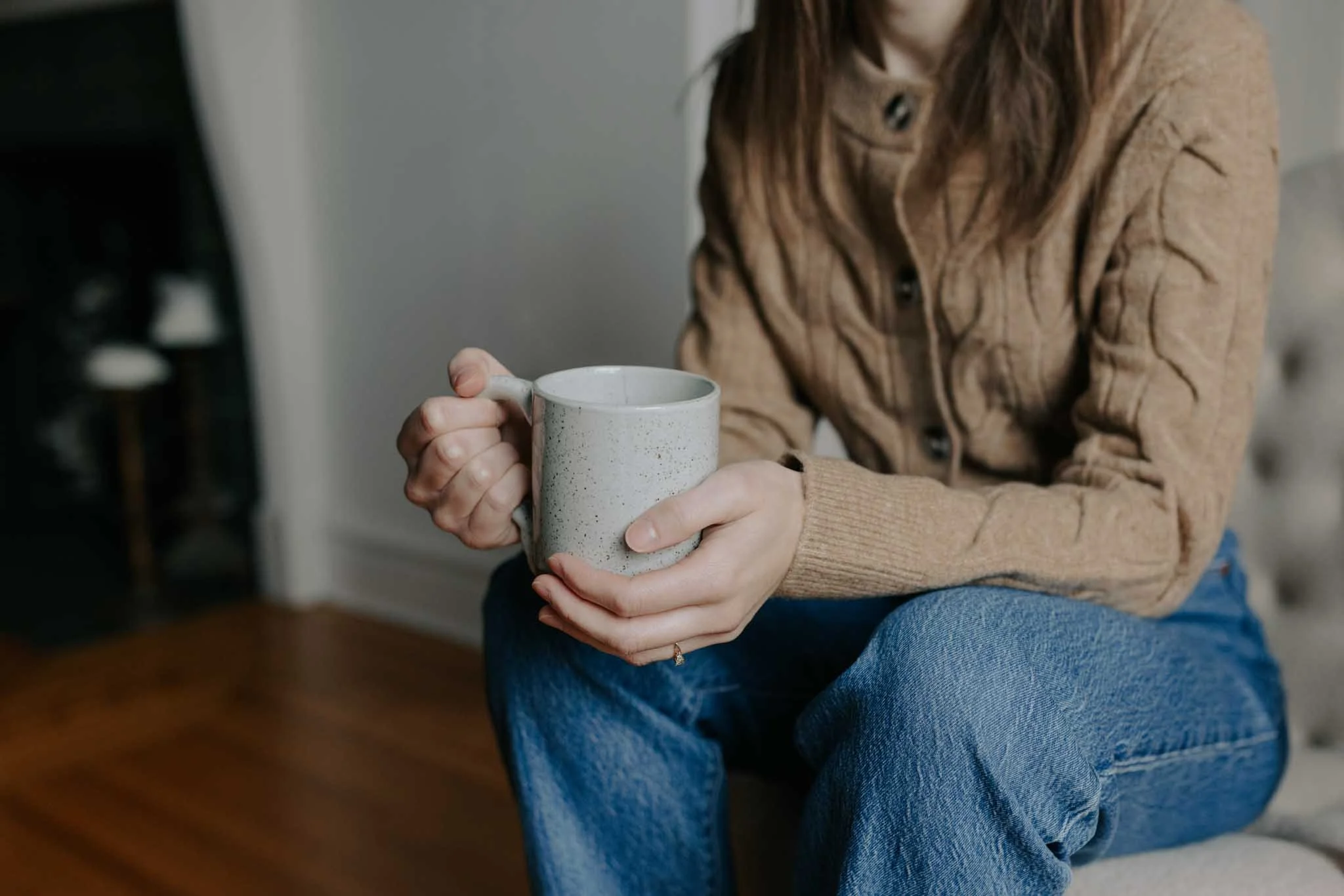 A woman wearing a tan cable knit sweater and blue jeans sitting indoors, holding a speckled ceramic mug with both hands.