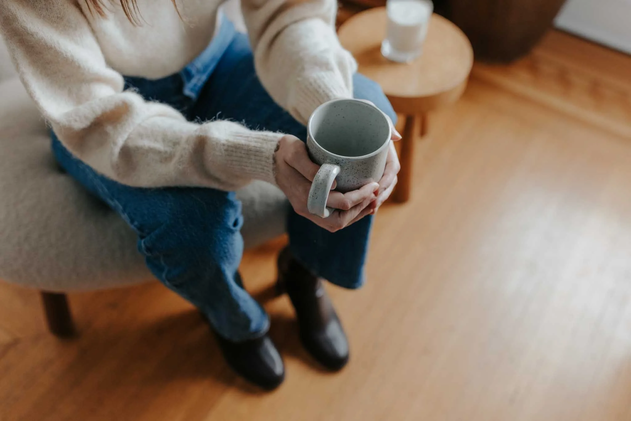 Person sitting on a beige cushioned chair holding a speckled ceramic mug, wearing a cream-colored sweater, jeans, and black shoes, next to a round wooden table with two glasses of water.