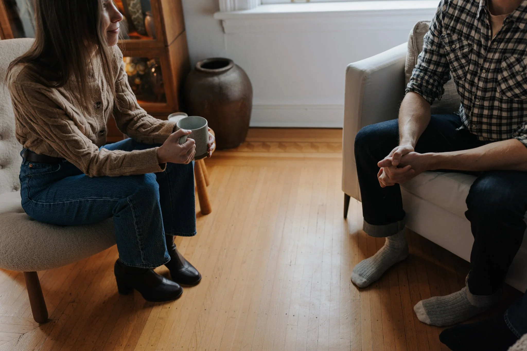 A woman sitting on a beige chair holding a mug, engaged in conversation with a man sitting on a sofa. The scene is in a cozy living room with wood flooring, a large ceramic vase, and wooden furniture.