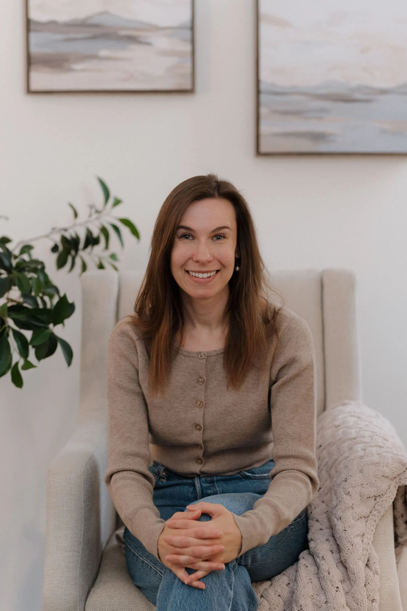 A woman with long brown hair, smiling, sitting on a beige couch in a room with abstract paintings and a plant. She's wearing a beige button-up sweater and blue jeans, with her hands clasped.