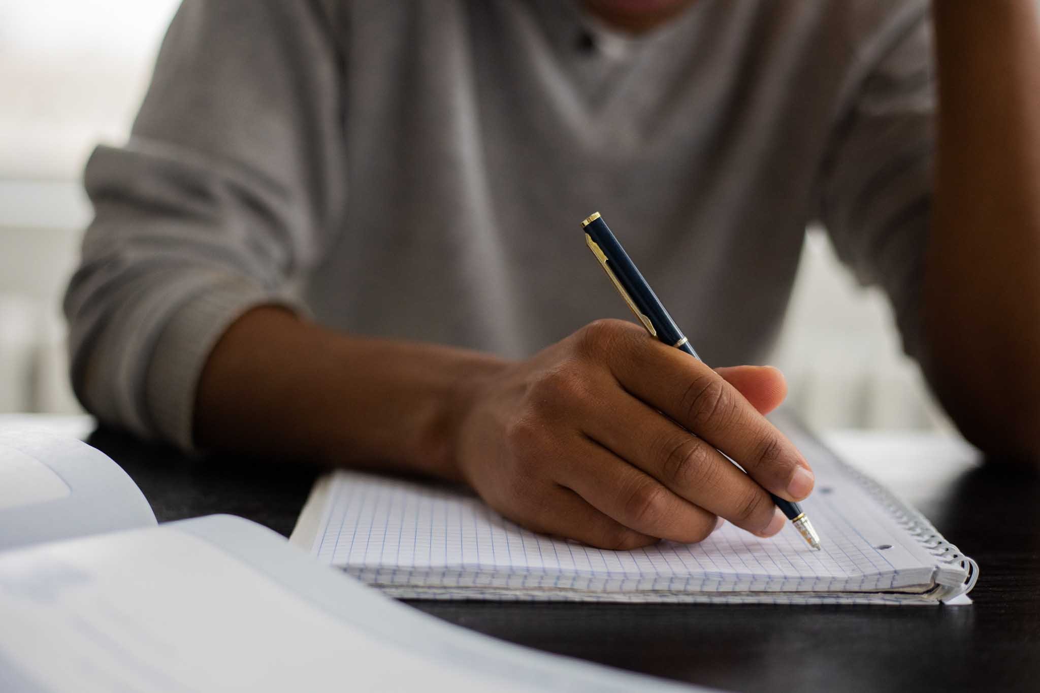 Person writing in a notebook with a pen, with a sheet of paper and a spiral notebook nearby.