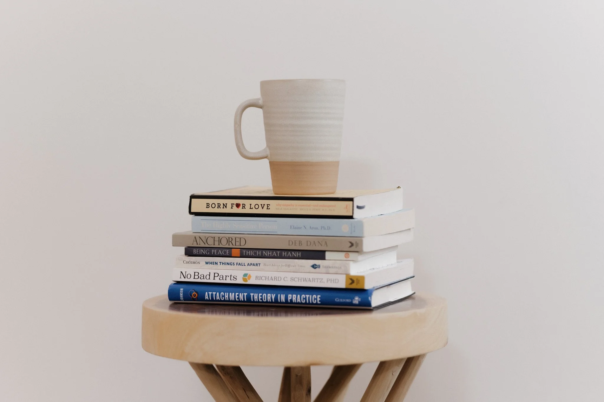 A wooden stool with a round top and crossed legs, holding a stack of ten books with a beige and white ceramic mug on top, against a plain white background.