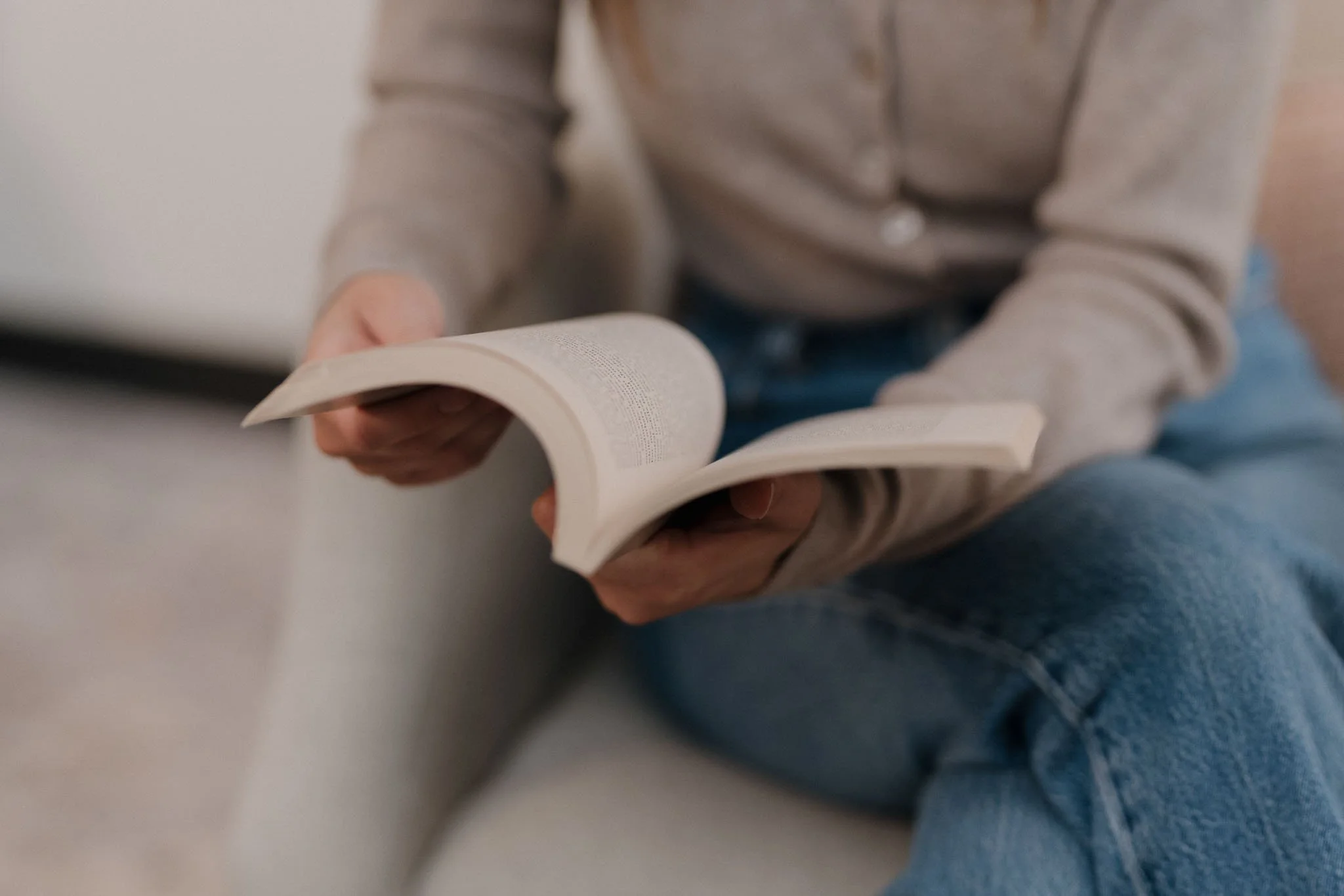 Person sitting on the floor reading a book, wearing a gray hoodie and blue jeans.