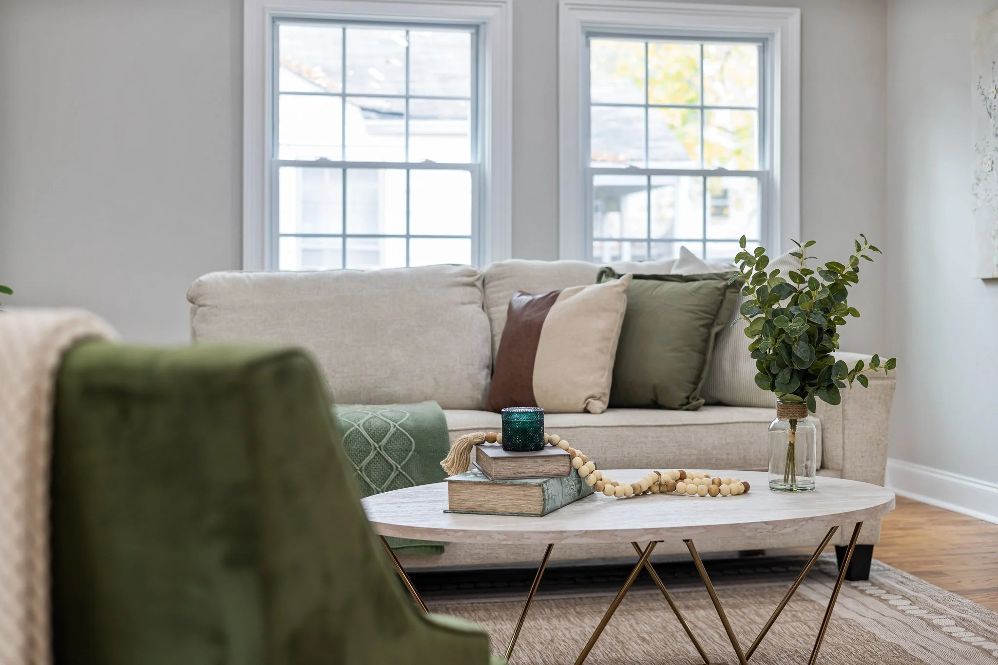 A cozy living room with a light-colored sofa adorned with multiple throw pillows, placed in front of two large windows. A wooden coffee table with decorative books, a candle, a beaded garland, and a glass vase with a leafy green plant adds to the inviting atmosphere. The room features hardwood floors and neutral wall colors, creating a bright and comfortable space.