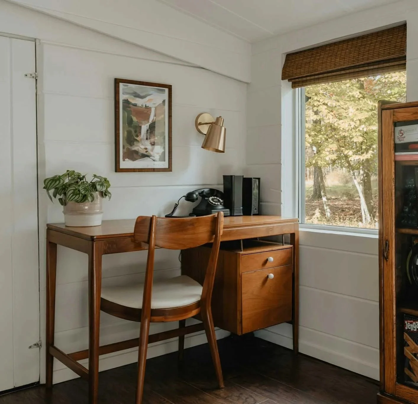 Interior of a cozy home office with a wooden desk, a vintage black rotary phone, a potted plant, and a window with brown blinds showing trees outside.