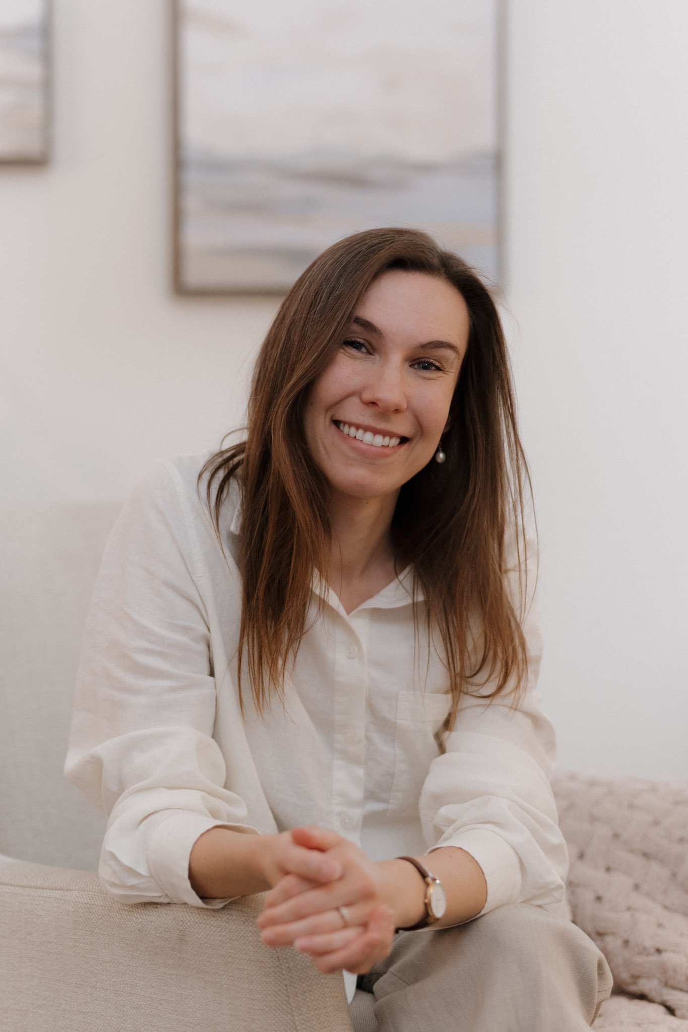A woman with long brown hair wearing a white shirt and a wristwatch, smiling and sitting on a beige couch with her hands clasped.