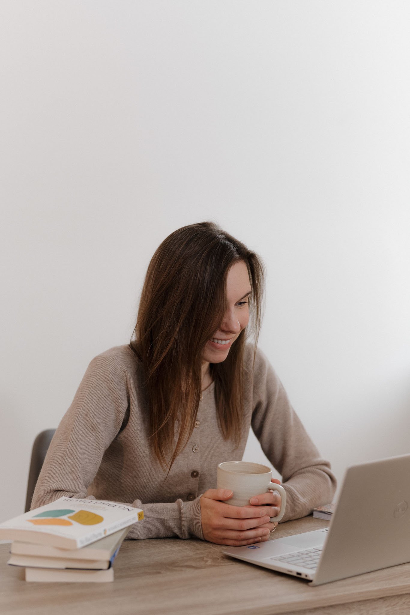 A woman with brown hair and a beige sweater sitting at a desk, holding a white mug, with books and a laptop in front of her.