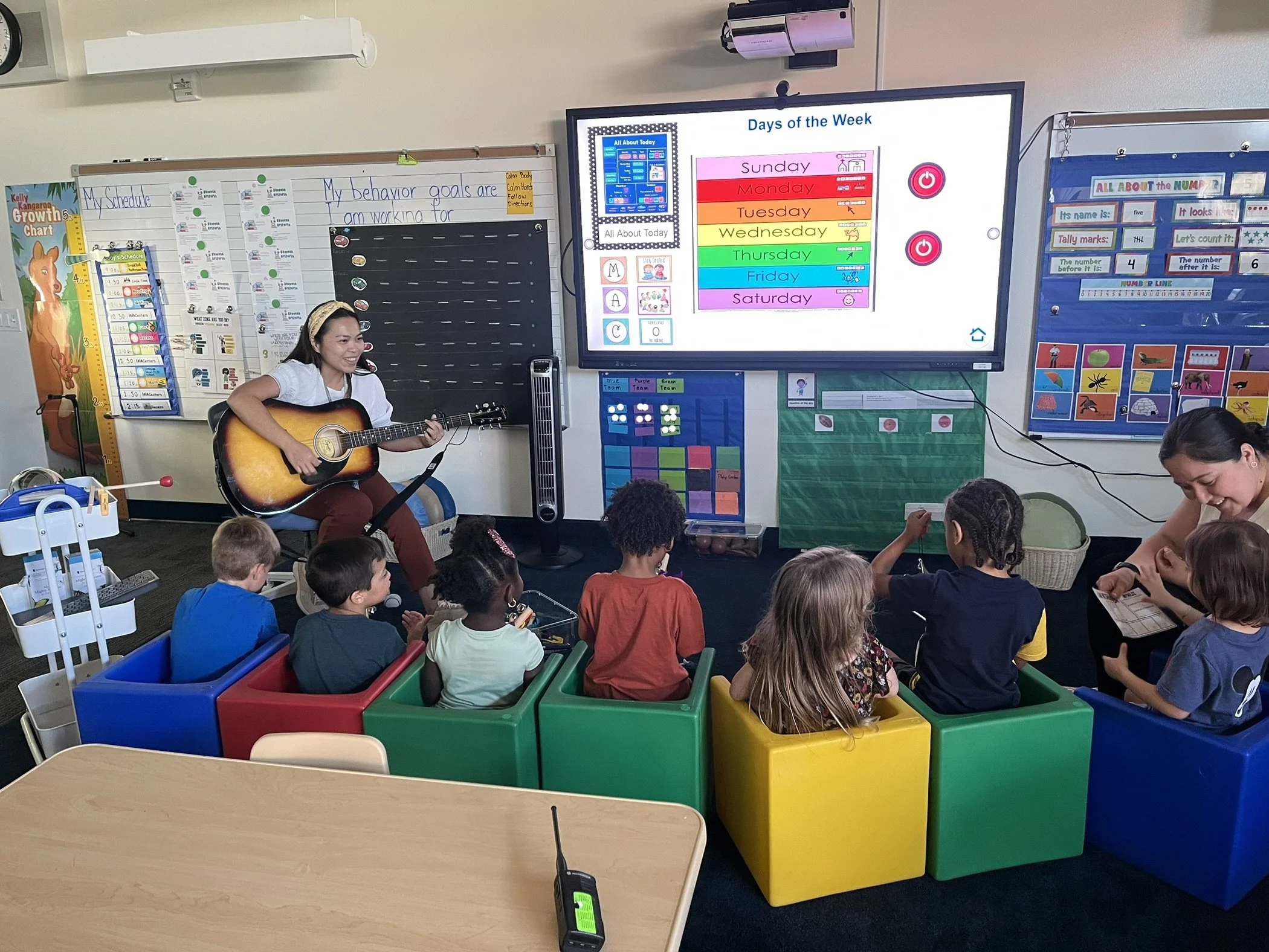 A classroom scene with a woman playing an acoustic guitar in front of a group of children. The classroom has colorful storage bins and educational posters, with a large digital screen displaying the days of the week.