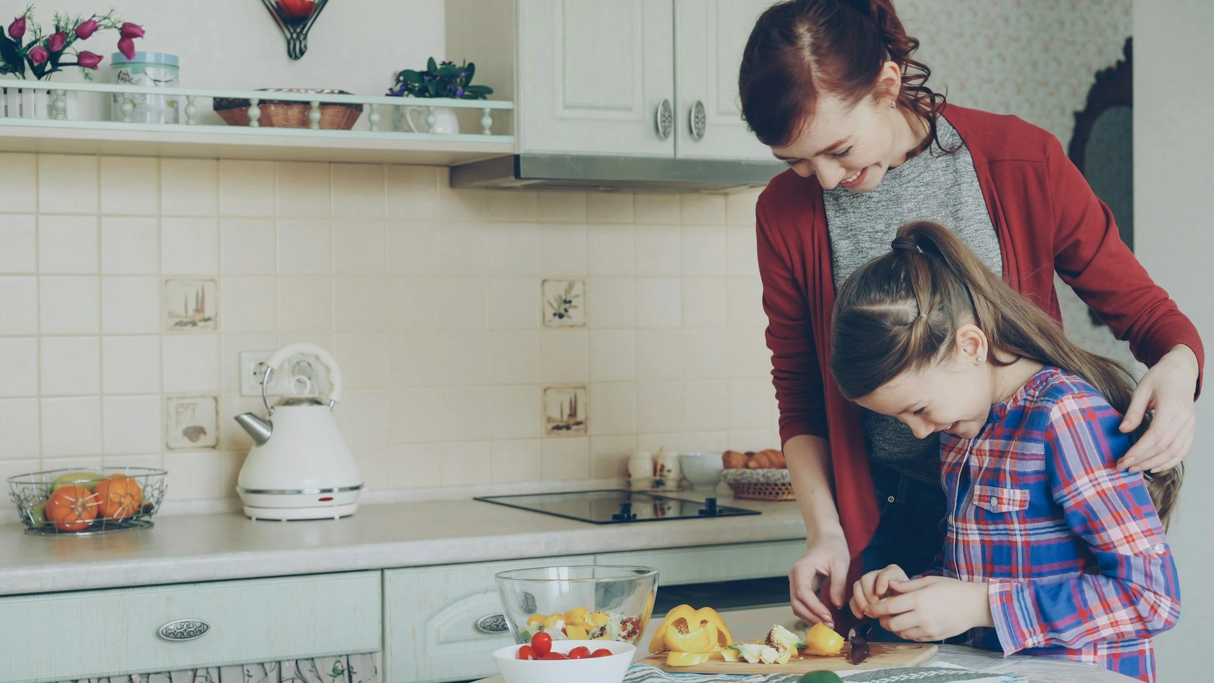 A woman with short red hair and a girl with long brown hair in a plaid shirt preparing food together in a kitchen, chopping ingredients on a cutting board.