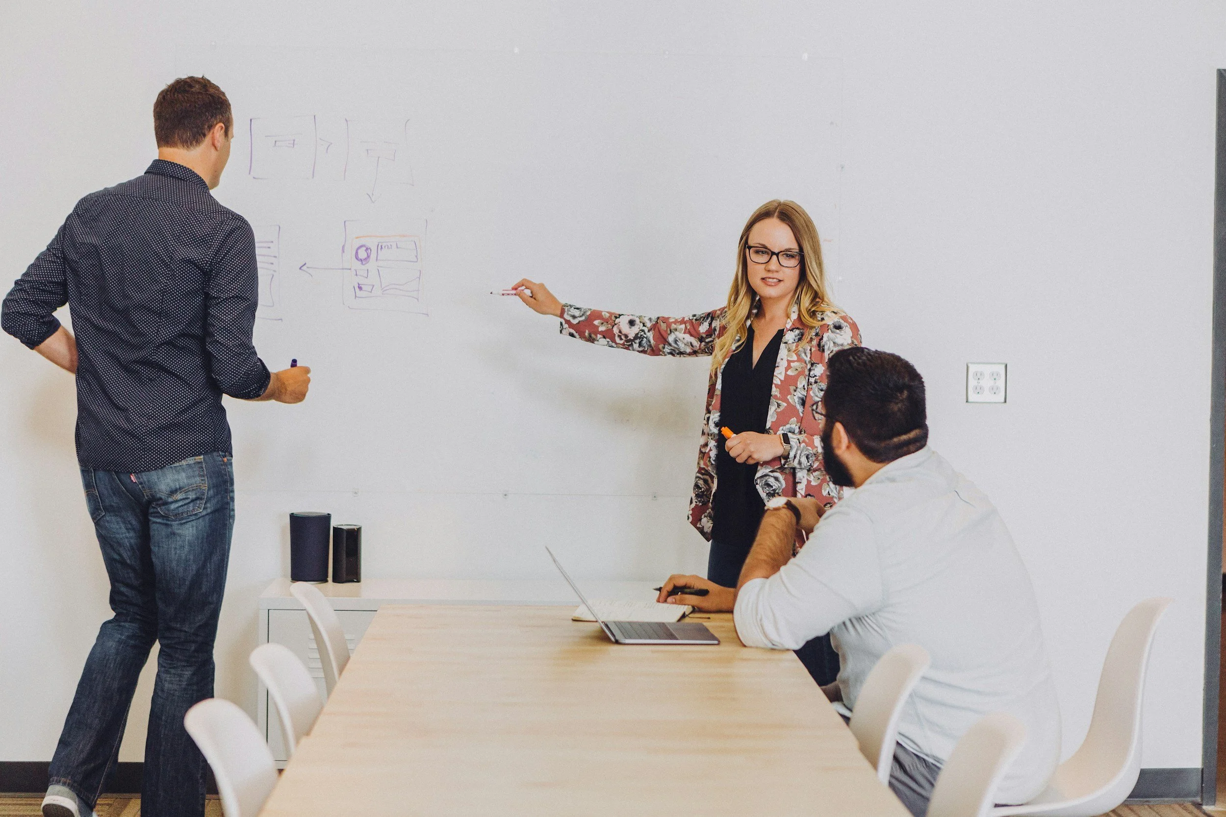 A group of four people engaged in a discussion in a modern office. One woman is standing by a whiteboard, pointing at it, while the others are seated at a desk with a laptop. The whiteboard contains diagrams and notes.