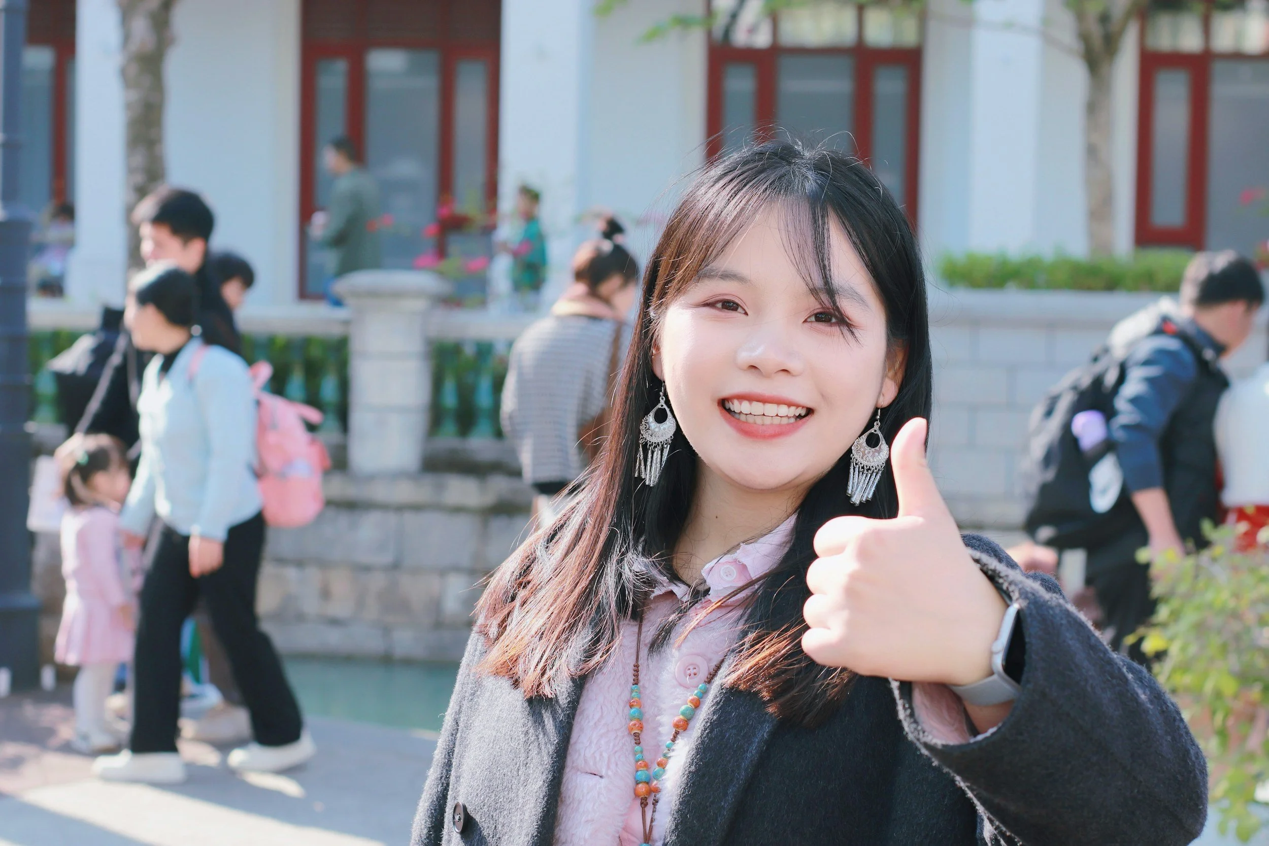 A young woman with black hair and earrings smiling and giving a thumbs-up gesture outdoors, with a group of people and a building in the background.