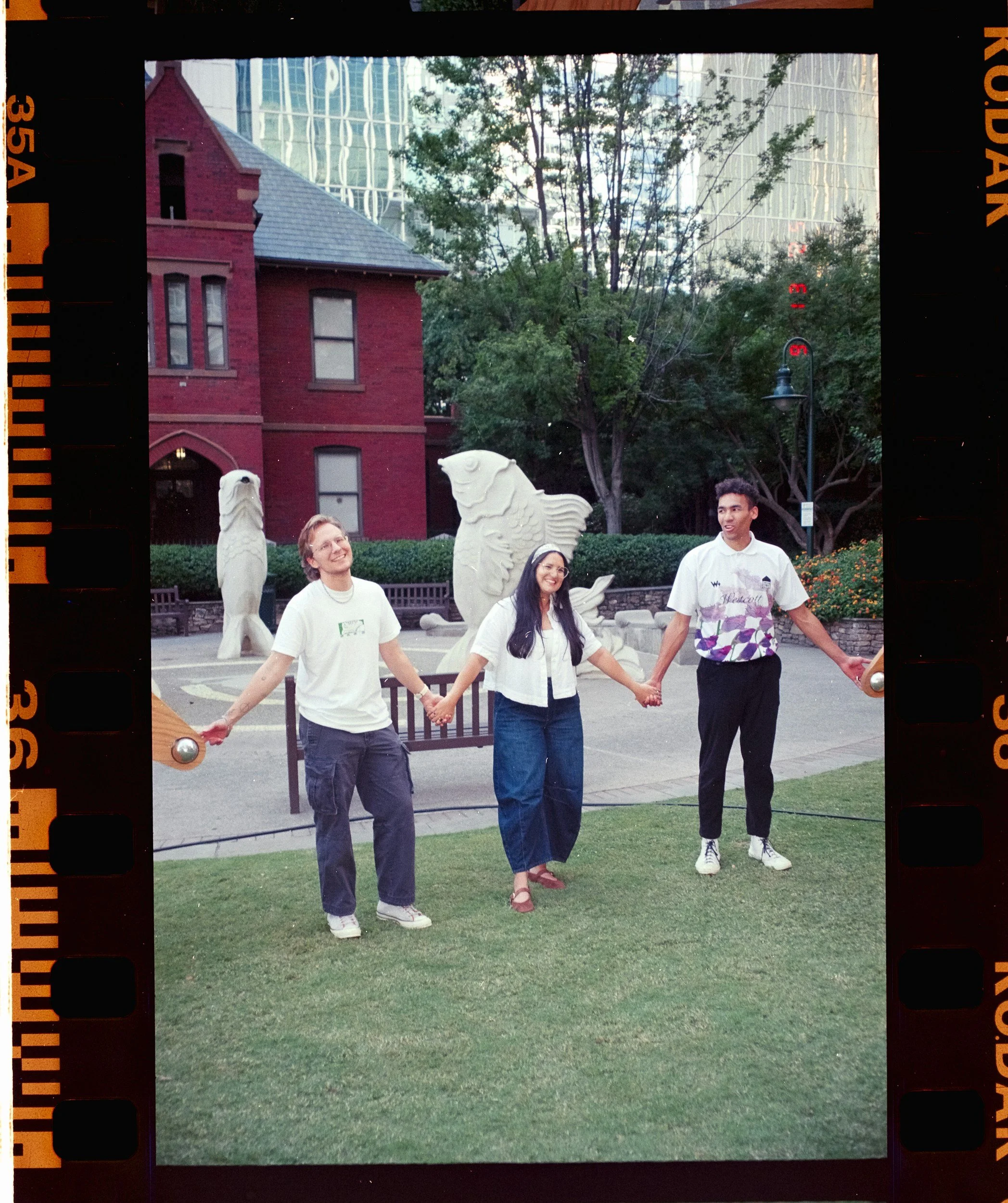 Four people hold hands in a circle outside on a grassy area, with a red brick building, trees, and modern glass buildings in the background.