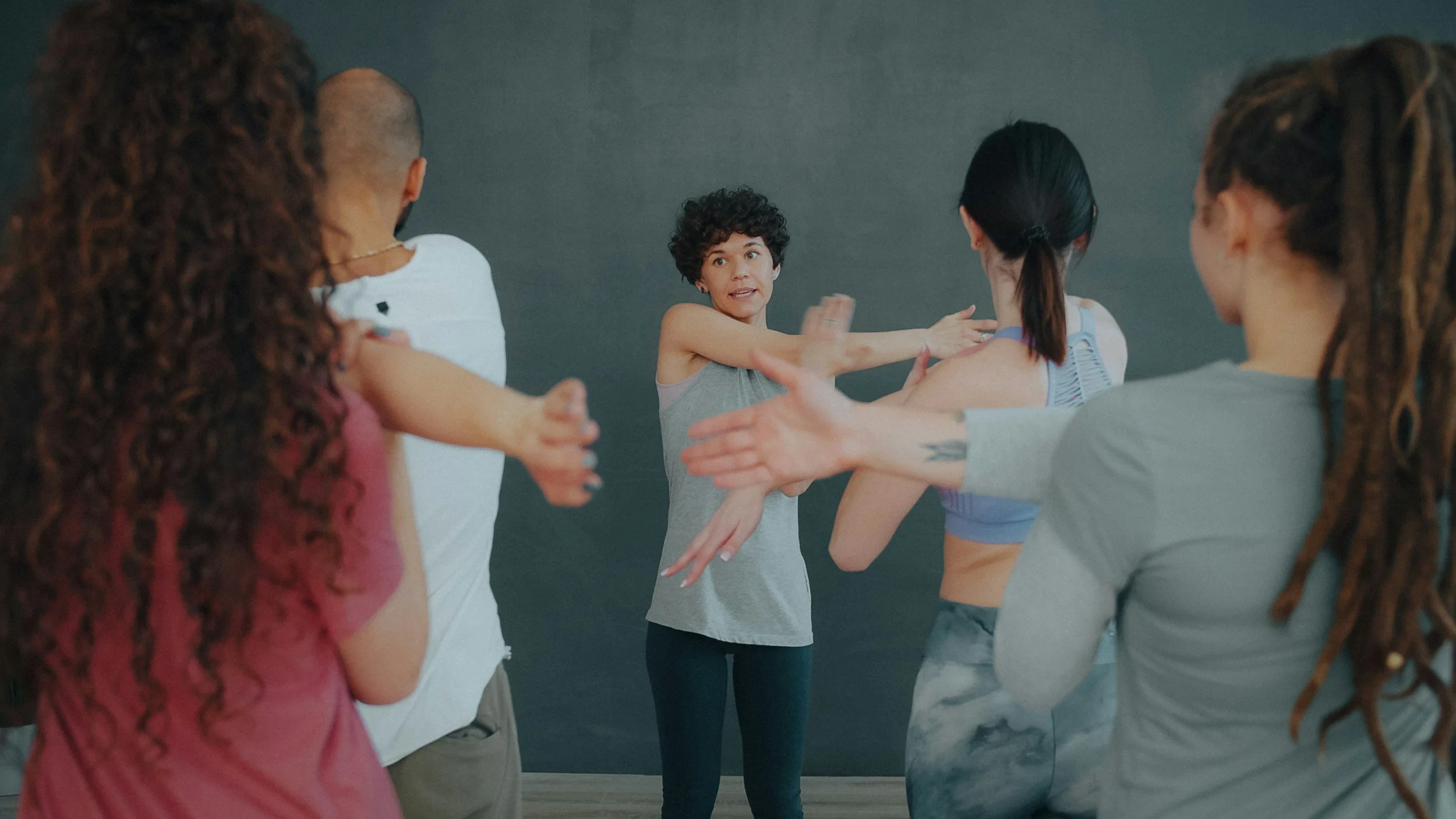 A fitness instructor demonstrating stretching exercises to a diverse group of women in a studio with gray walls.