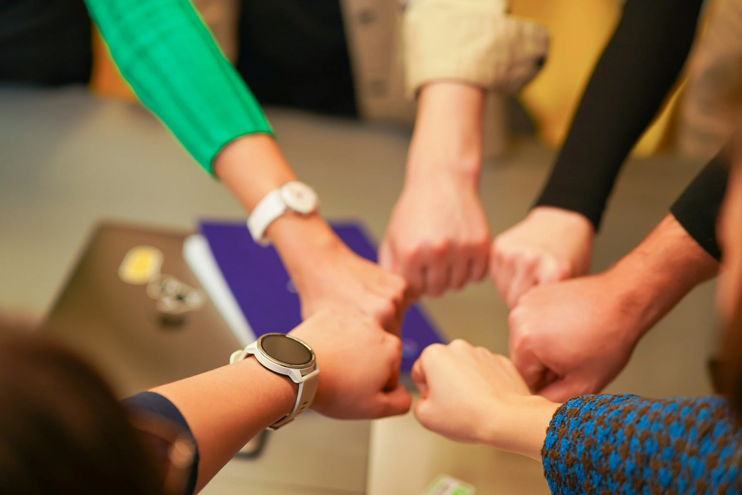 Several people combined hands in a gesture of unity or teamwork, with some individuals wearing watches, bracelets, and colorful clothing.