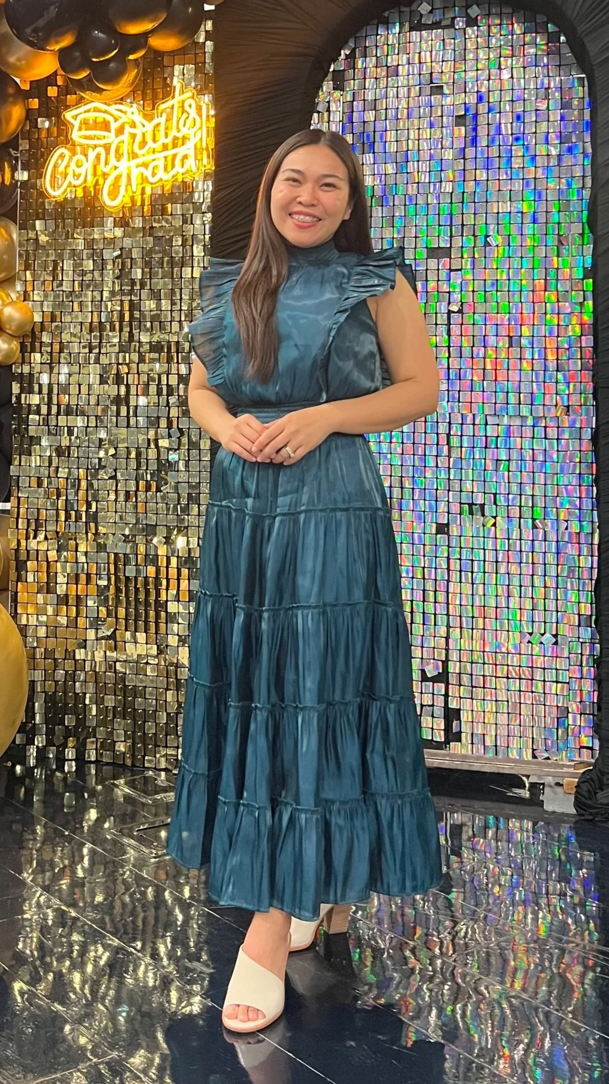 A woman in a tiered blue dress standing in front of a holographic mosaic wall with a neon sign that reads 'Birthday Goals' surrounded by balloons.