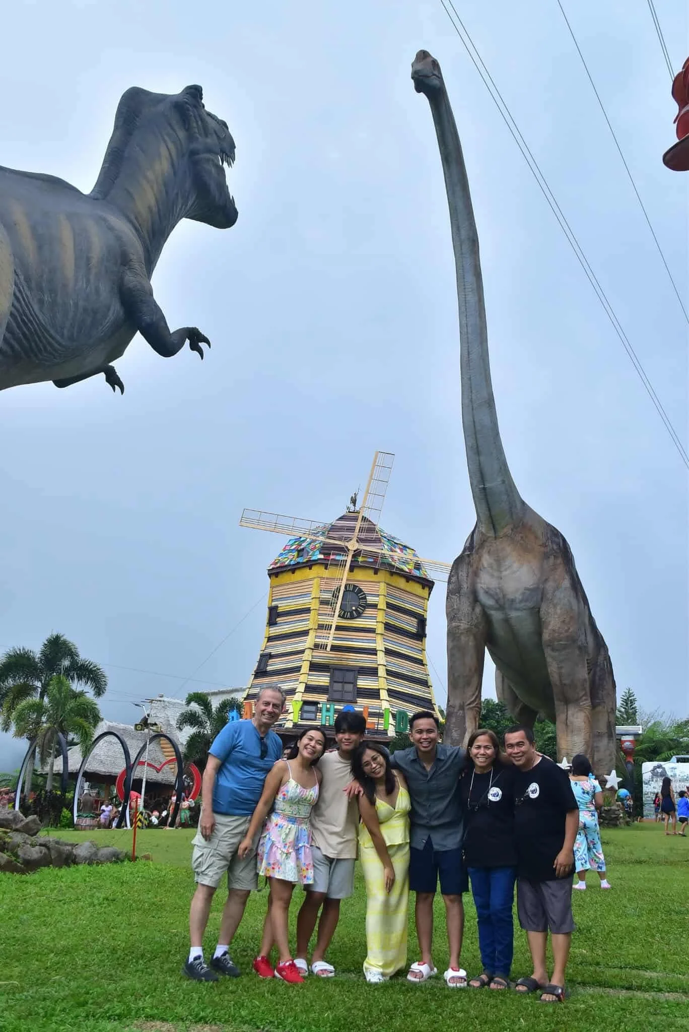 Group of seven people standing on grass in front of a colorful windmill with a clock, two tall dinosaur statues, and a theme park setting.