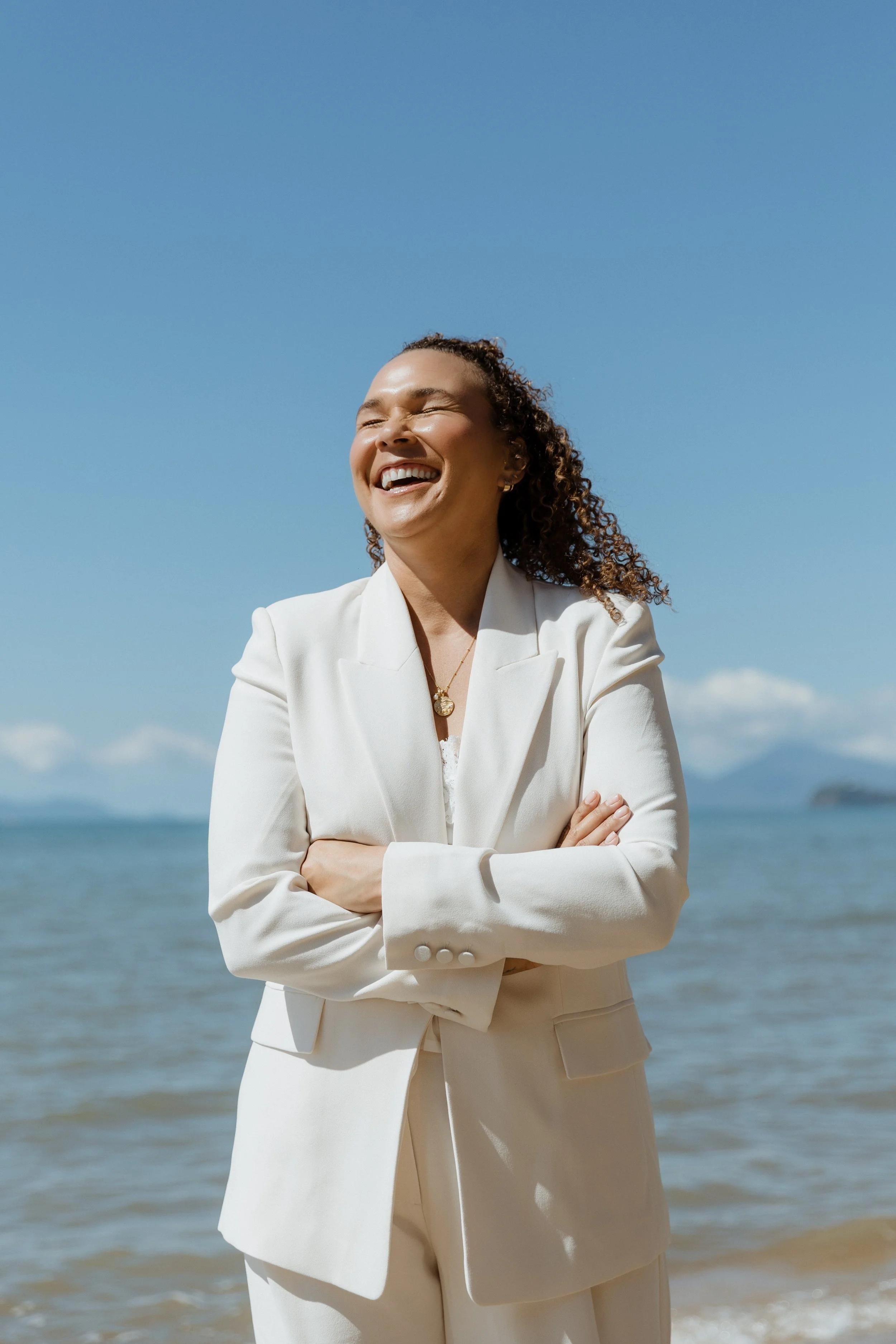 A woman in a white suit smiling and laughing with arms crossed, standing on a beach with the ocean and a blue sky in the background.