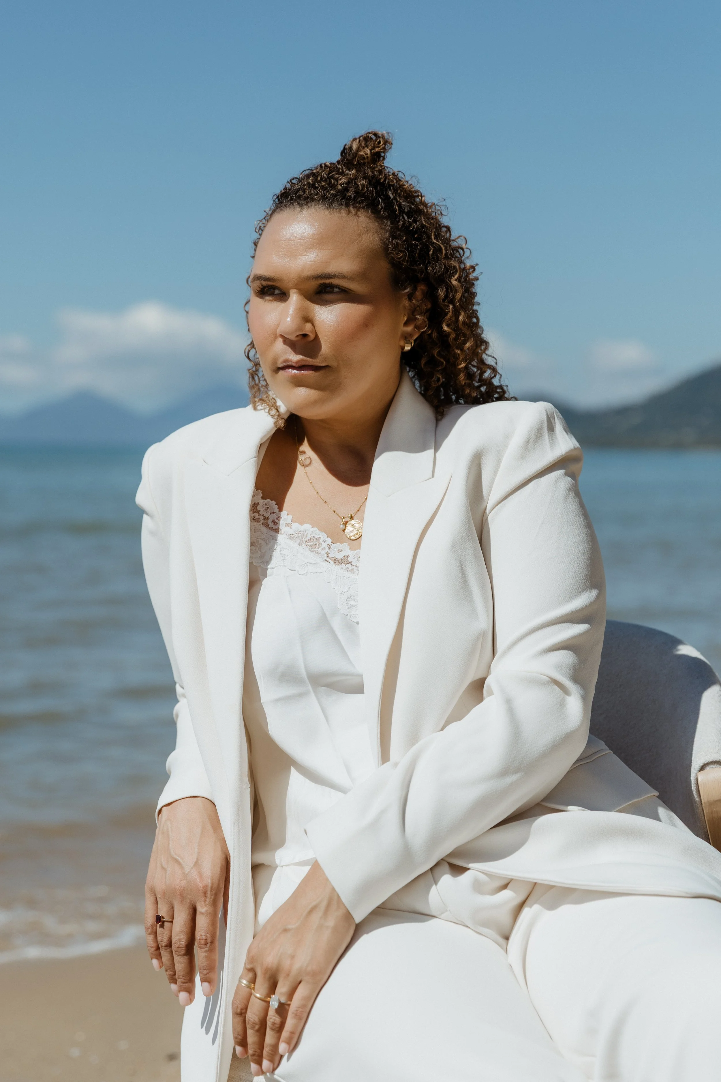A woman with curly hair wearing a white blazer and pants sitting outdoors near a body of water, with mountains and a blue sky in the background.