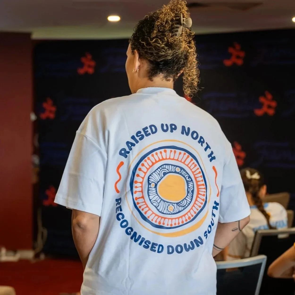 Back view of a person wearing a white t-shirt with a circular logo and text that reads "Raised Up North Recognised Down South" at an indoor event with other attendees in the background.