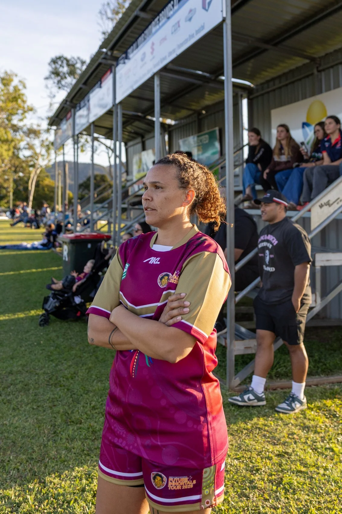 A female rugby player stands with her arms crossed, wearing a maroon and gold uniform, at a sports event on a grassy field with a spectator stand in the background. Other spectators are seated on the stand.