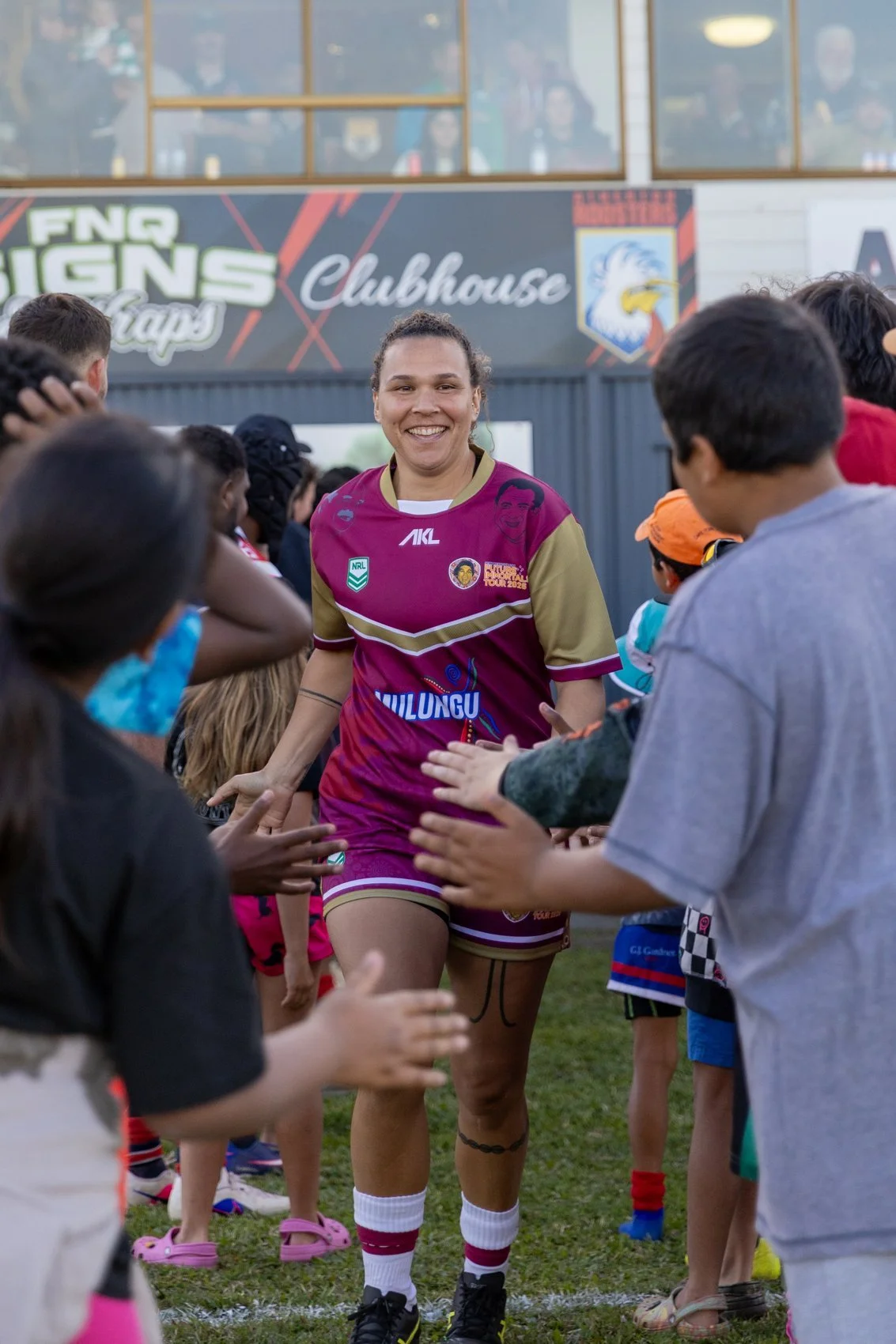 A female rugby player in a maroon and gold uniform walking through a crowd of children on a rugby field, smiling. The children are reaching out to her, and some are wearing face masks. In the background, there is a sign that reads 'FNC Signs Clubhouse' and an area with onlookers.
