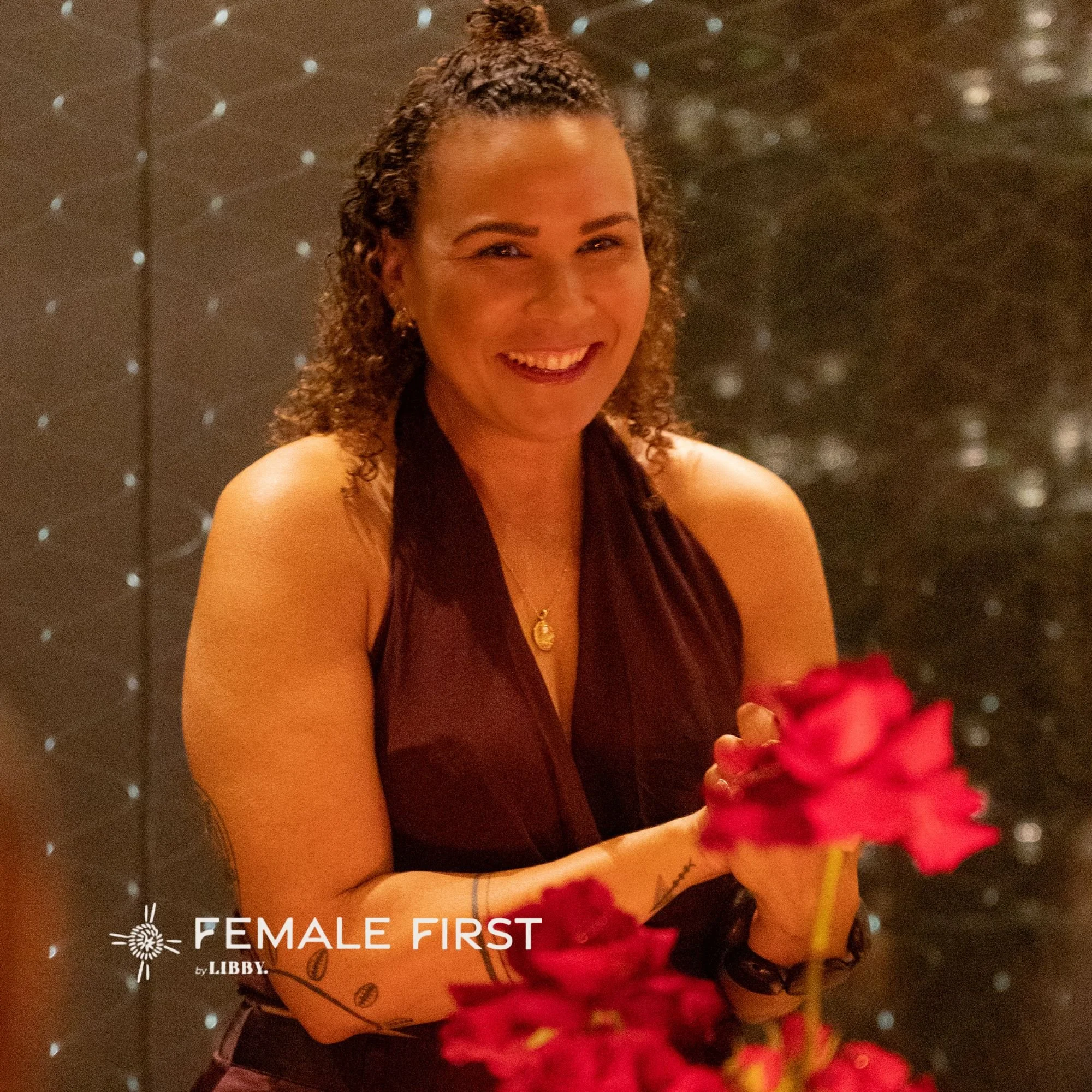 A smiling woman with curly hair, wearing a sleeveless top and jewelry, holding a bouquet of red flowers in front of a dark, sparkly background.