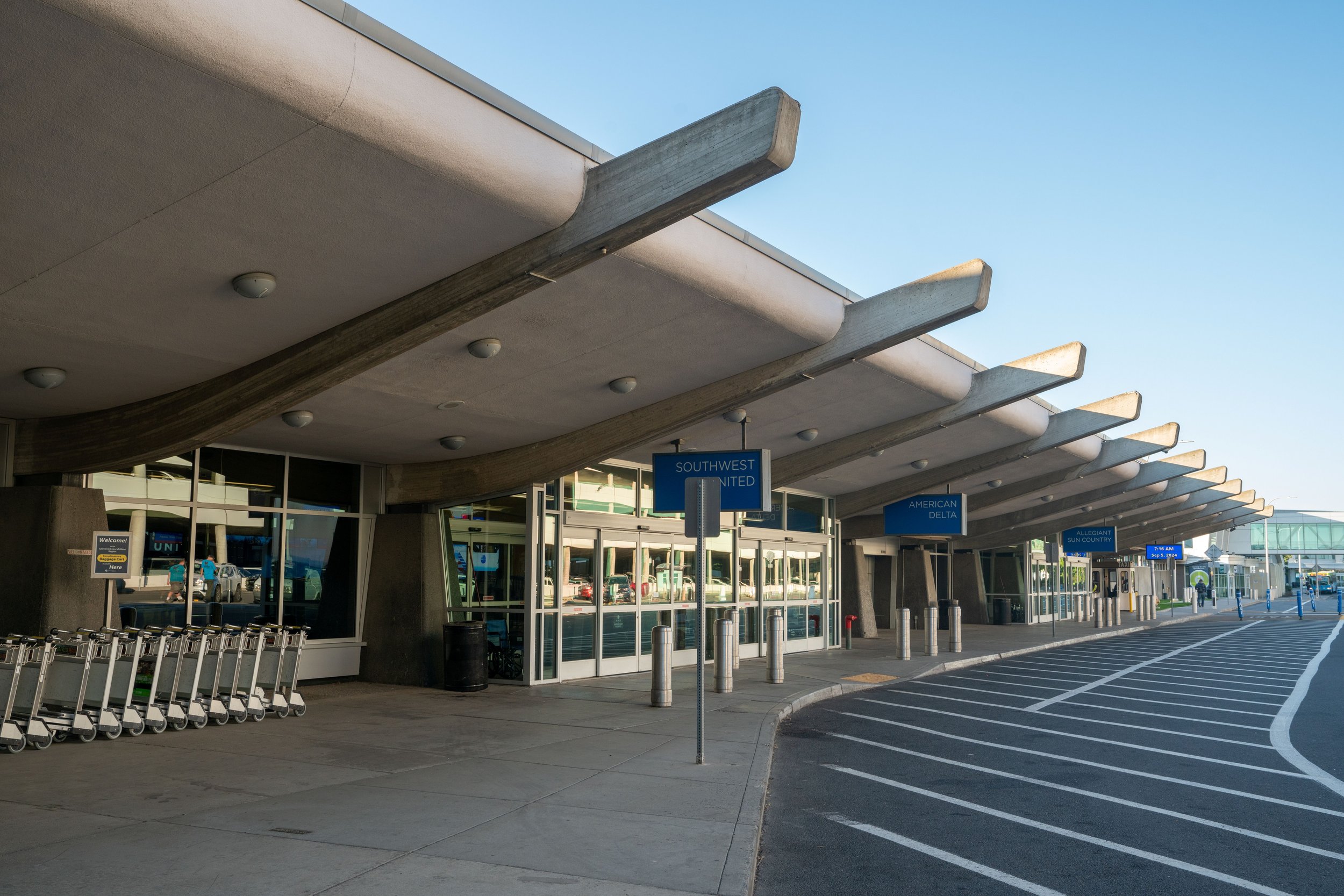 Exterior view of an airport terminal with a modern architectural design featuring curved concrete and glass elements, airline signage, a row of luggage carts, and a few people walking outside on a clear day.