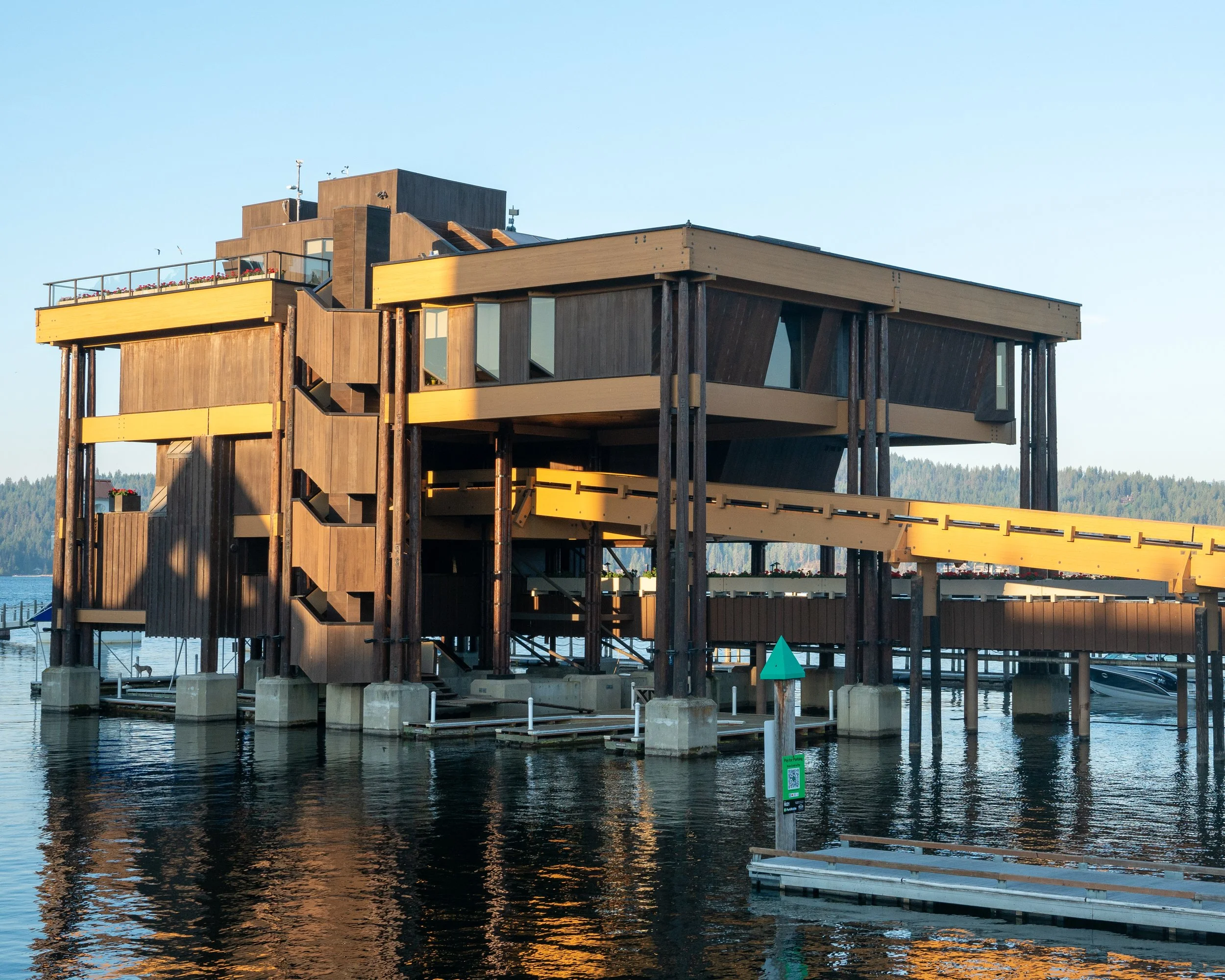 A modern wooden building on stilts over water, with a yellow accent, multiple balconies, and large windows, likely a waterfront house or lodge, with sailboats and dock in the background under a clear sky.