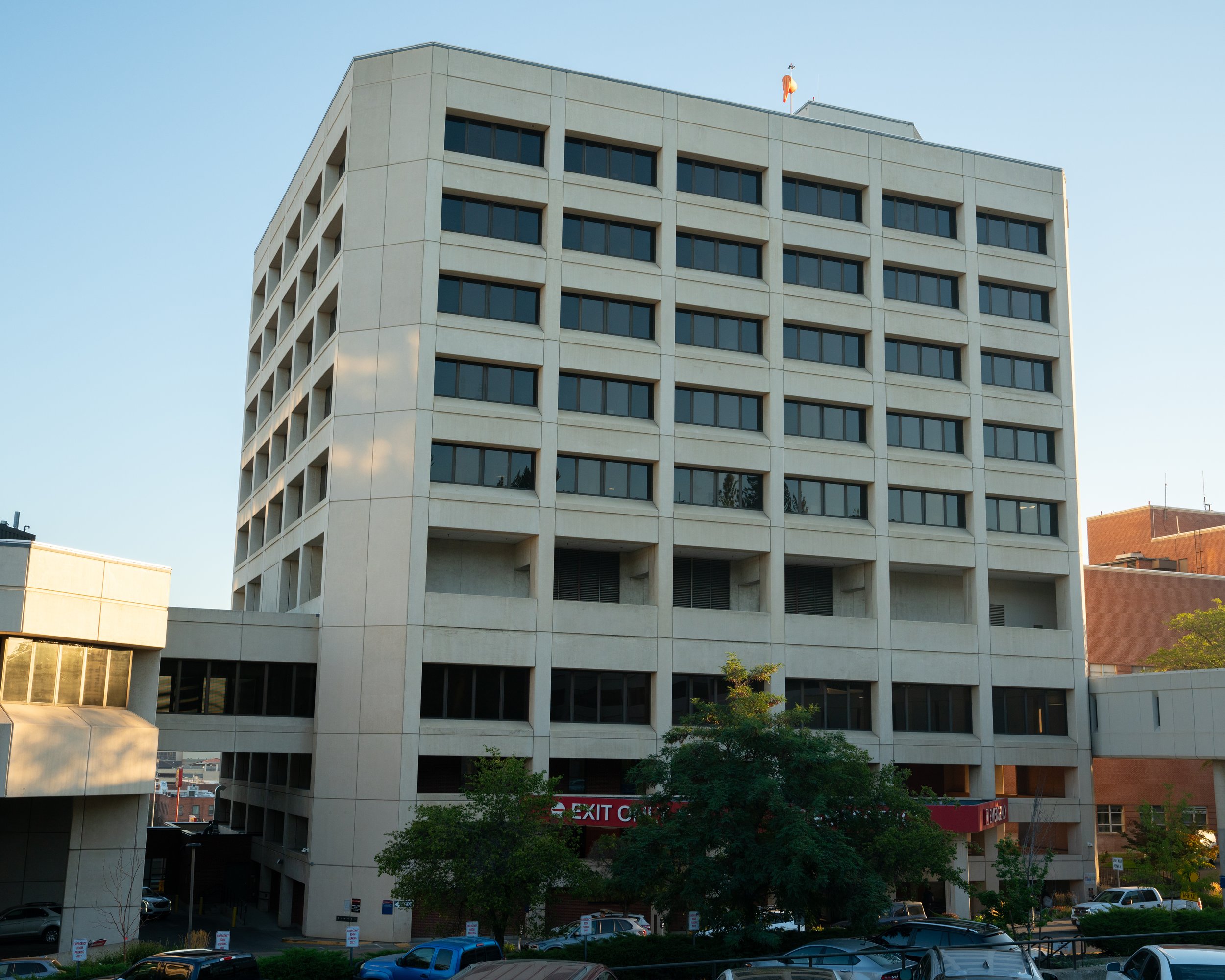 A tall, modern multi-story office building with rectangular windows and a curved corner. The street in front has parked cars, and a sign reads 'EXIT' on the lower level. Trees and a clear blue sky are visible.