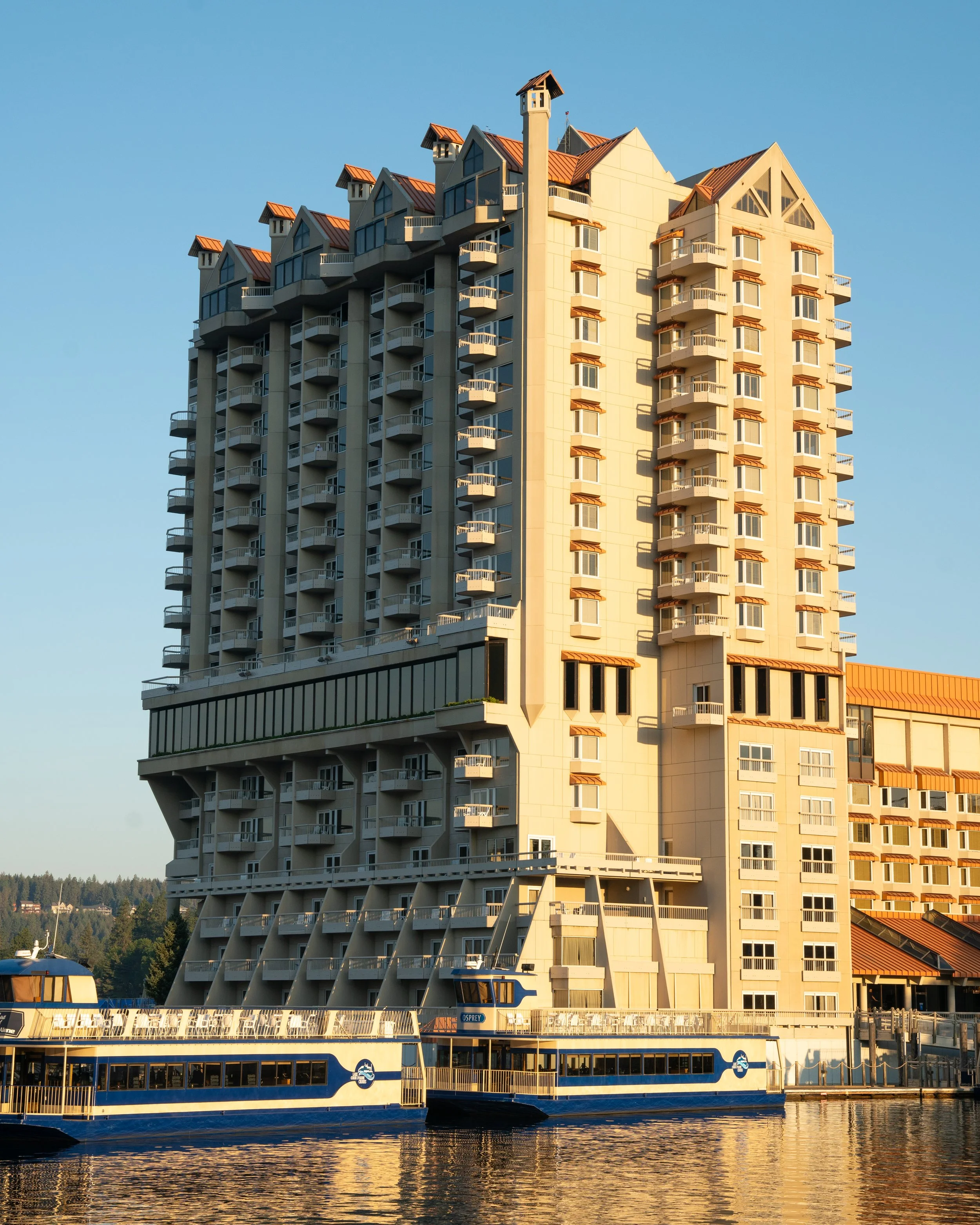 A large modern multi-story building with numerous balconies stands by the water, with a blue boat docked at a pier in the foreground. The sky is clear and blue.